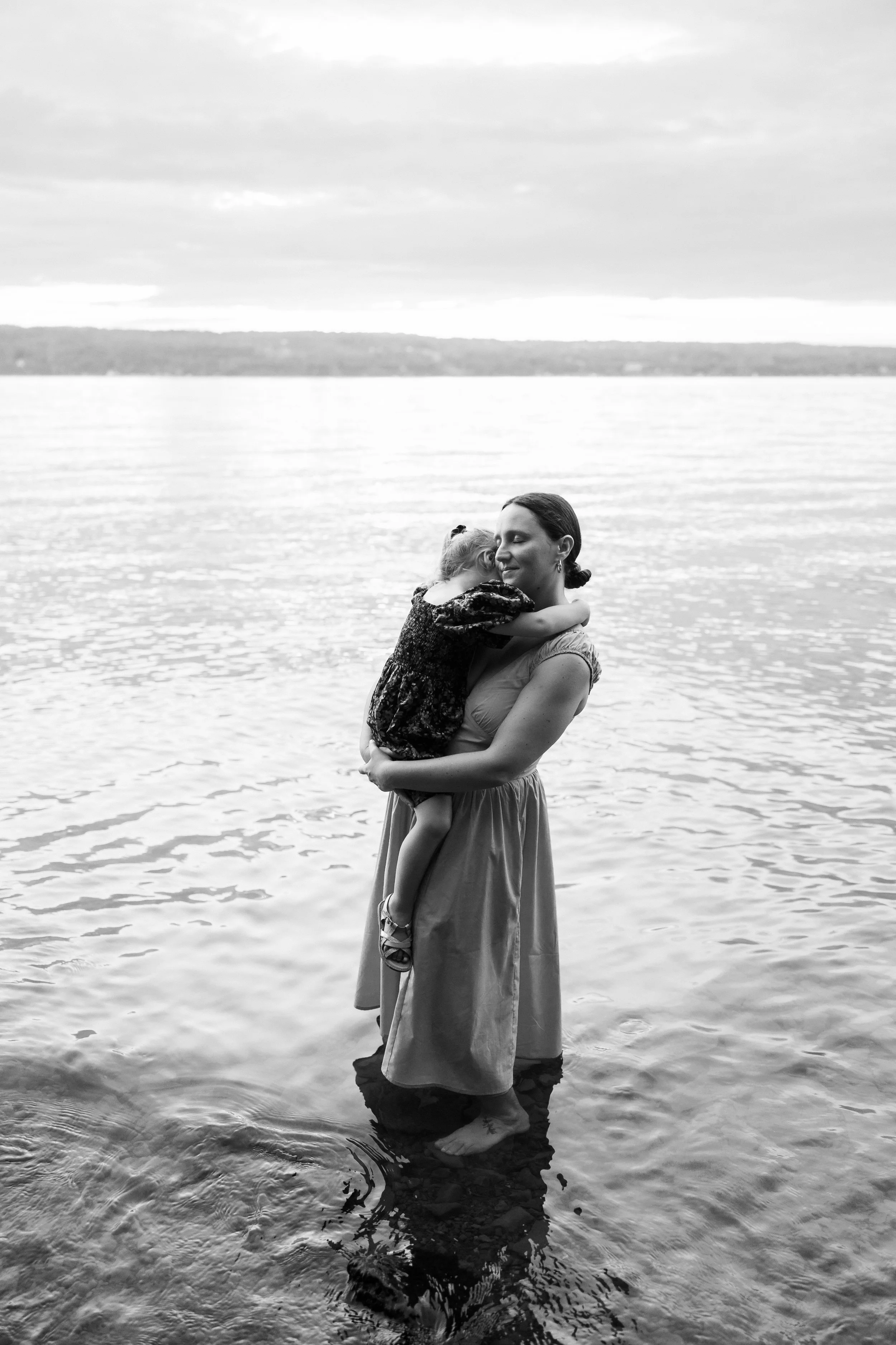A woman standing barefoot in water, holding her daughter in her arms, embracing in Seneca Lake.