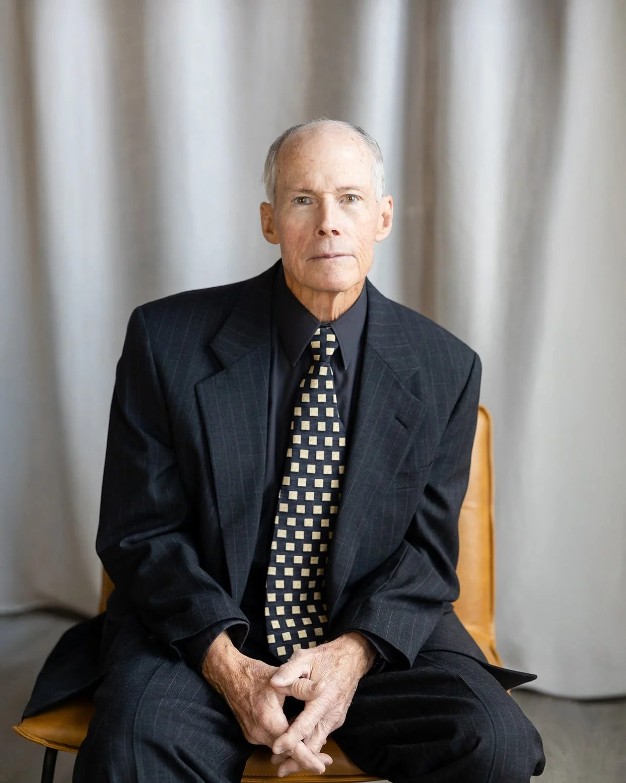 An elderly man with a serious expression, seated on a chair in front of a neutral background, wearing a dark suit, black shirt, and patterned tie.