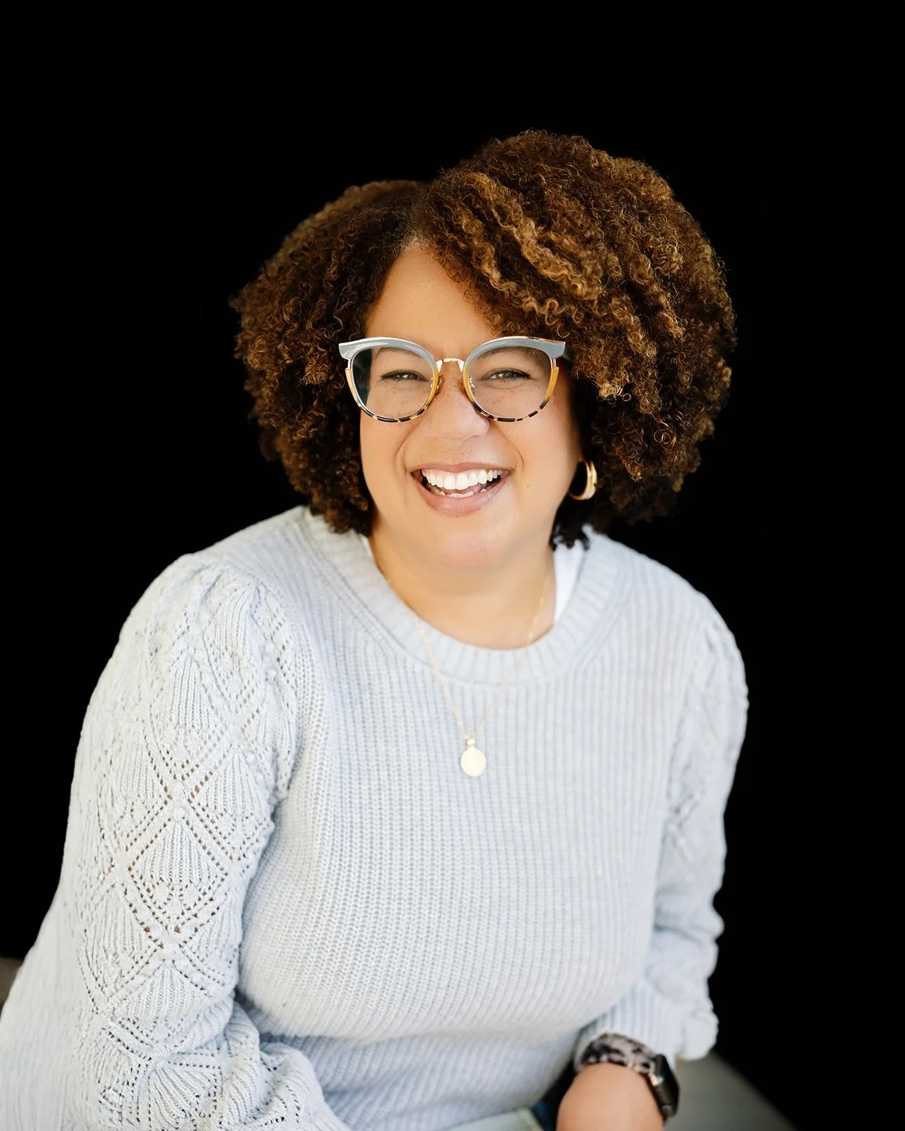 Professional headshot of a woman with wavy light brown hair, wearing a white shirt and light blue jeans, standing in a bright white Anderson Arts Building studio.