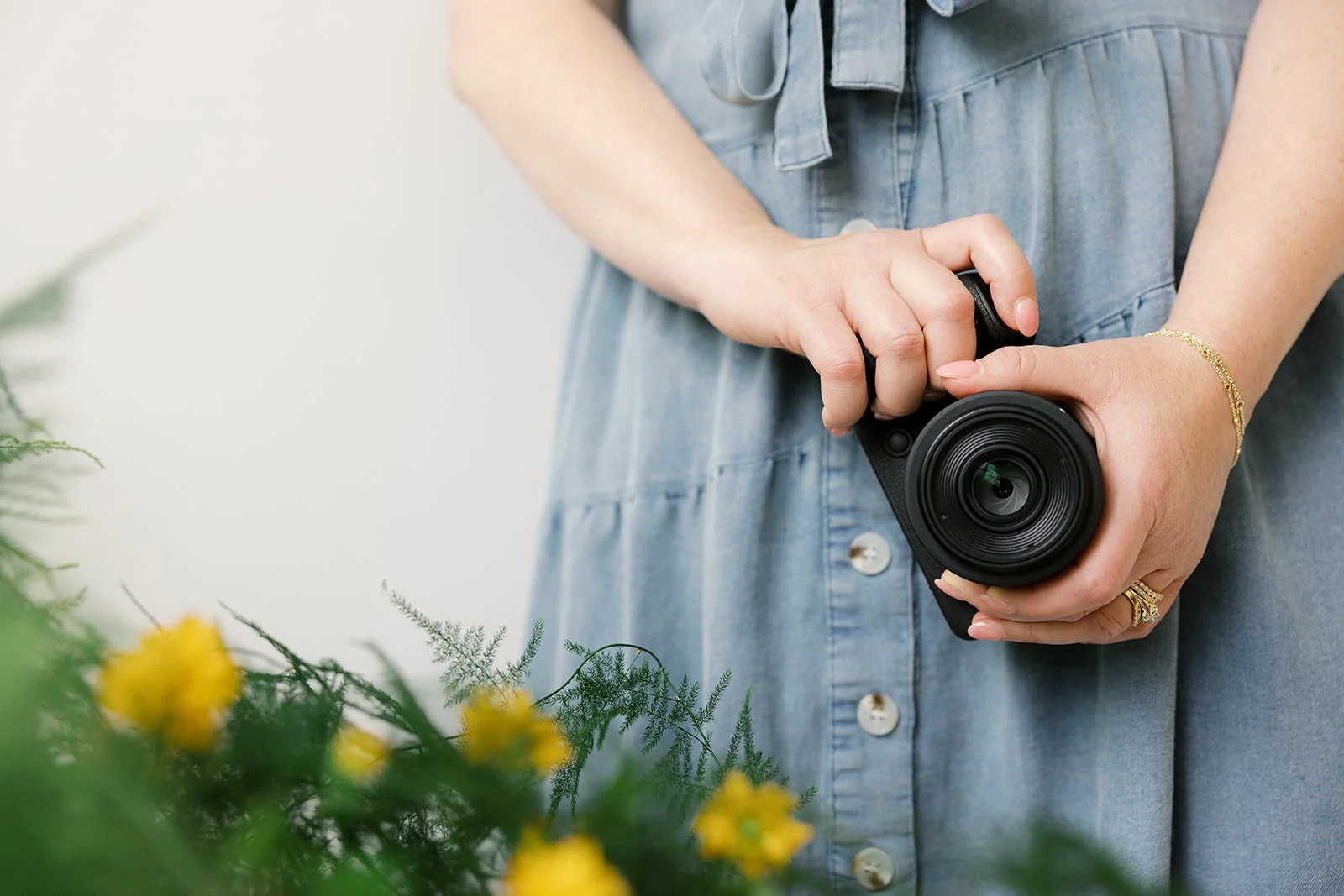 A woman holding a black camera with a large lens, wearing a blue dress with buttons, a gold bracelet, and a gold ring, with yellow flowers in the foreground.