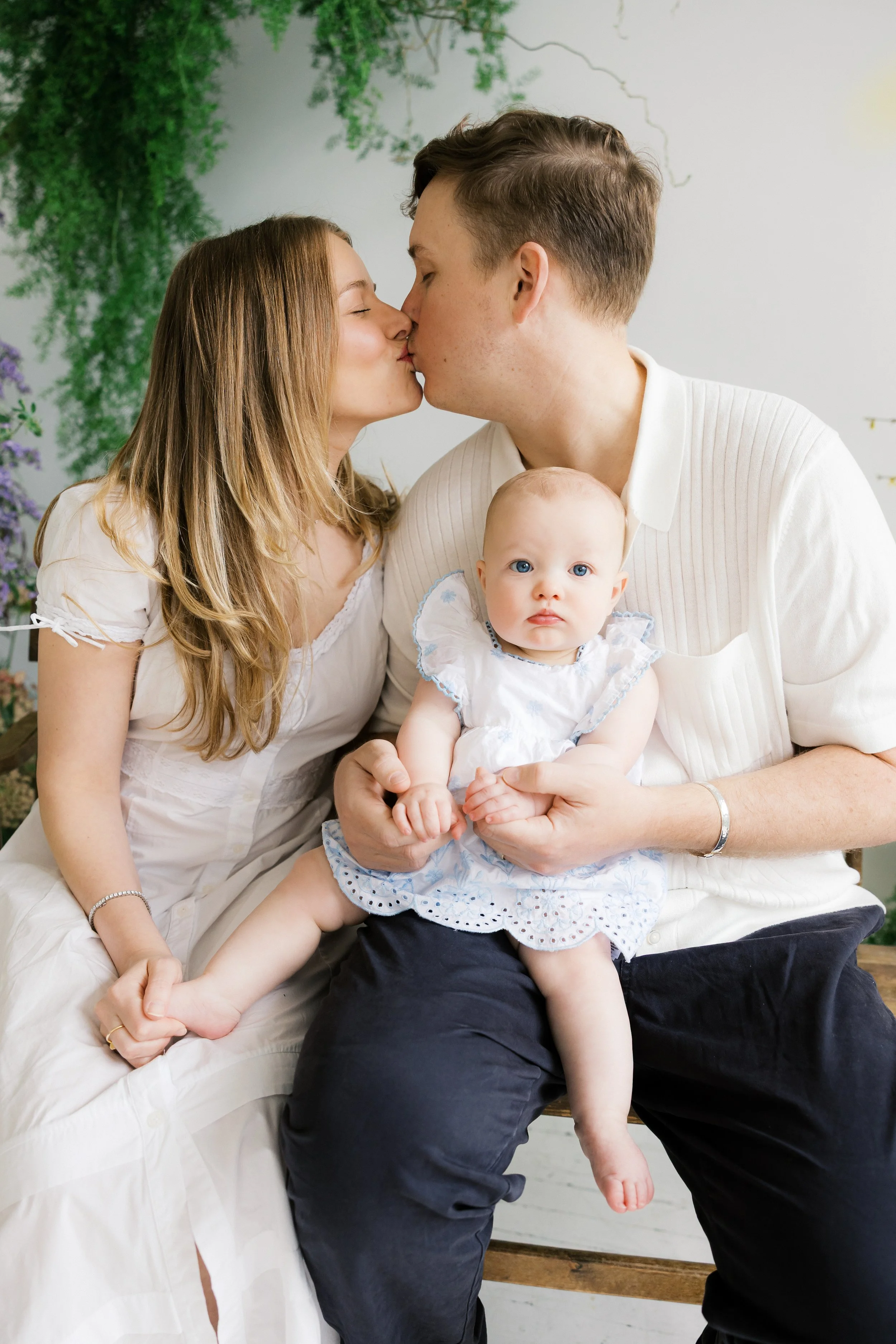 A couple is kissing while their baby girl sits on the father’s  lap during a studio family session in Rochester, NY.