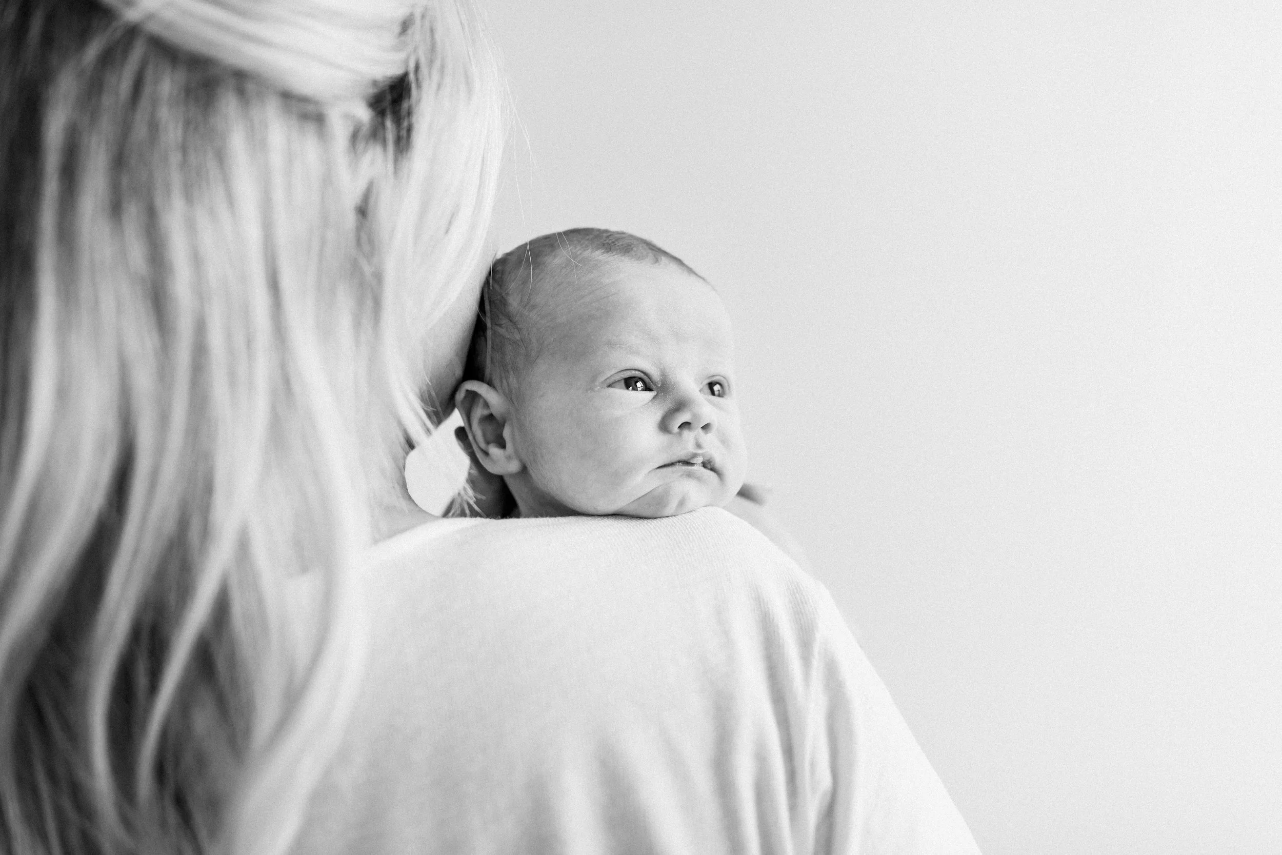 A woman with long blond hair holding a baby, who is resting on her shoulder and looking to the side, during an in-home newborn session in Rochester, NY.