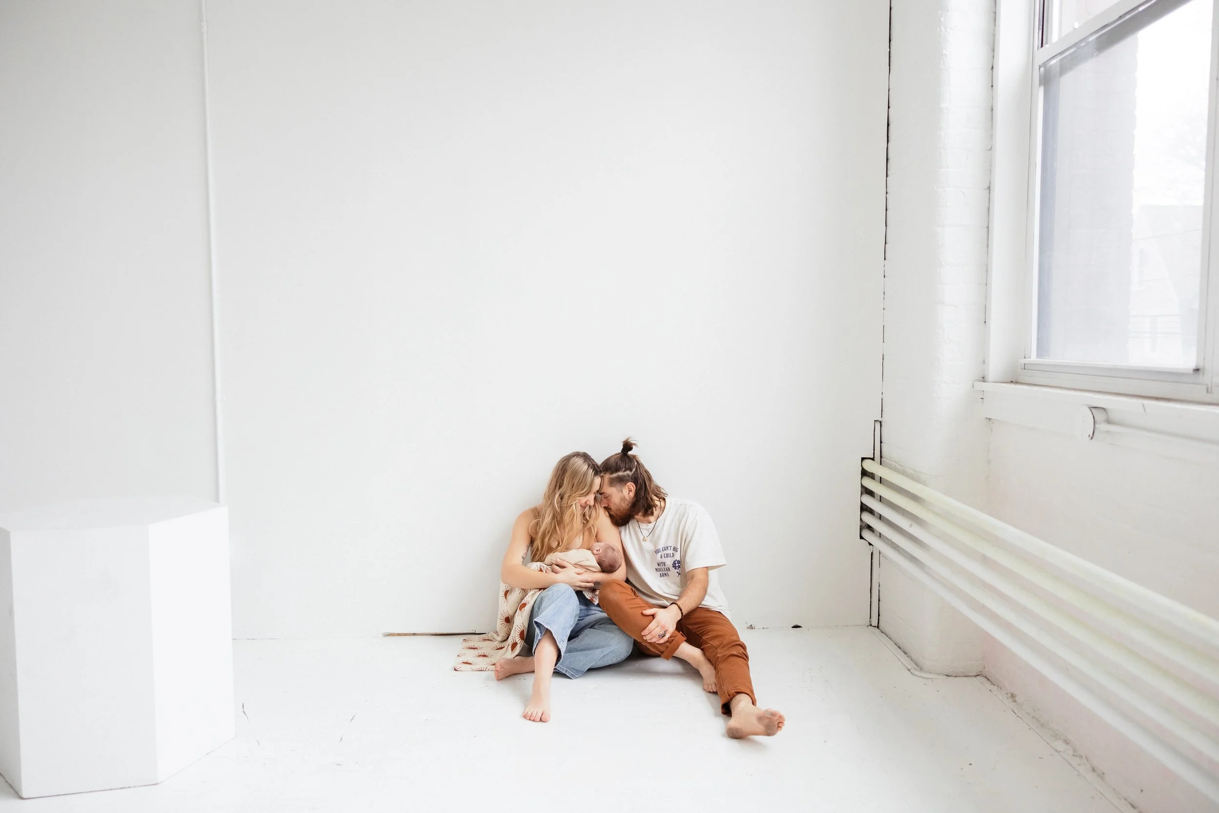 A couple sitting on a white floor in a minimalist room, tenderly holding and feeding their newborn baby during a studio newborn session in Rochester, NY at the Anderson Arts building. 