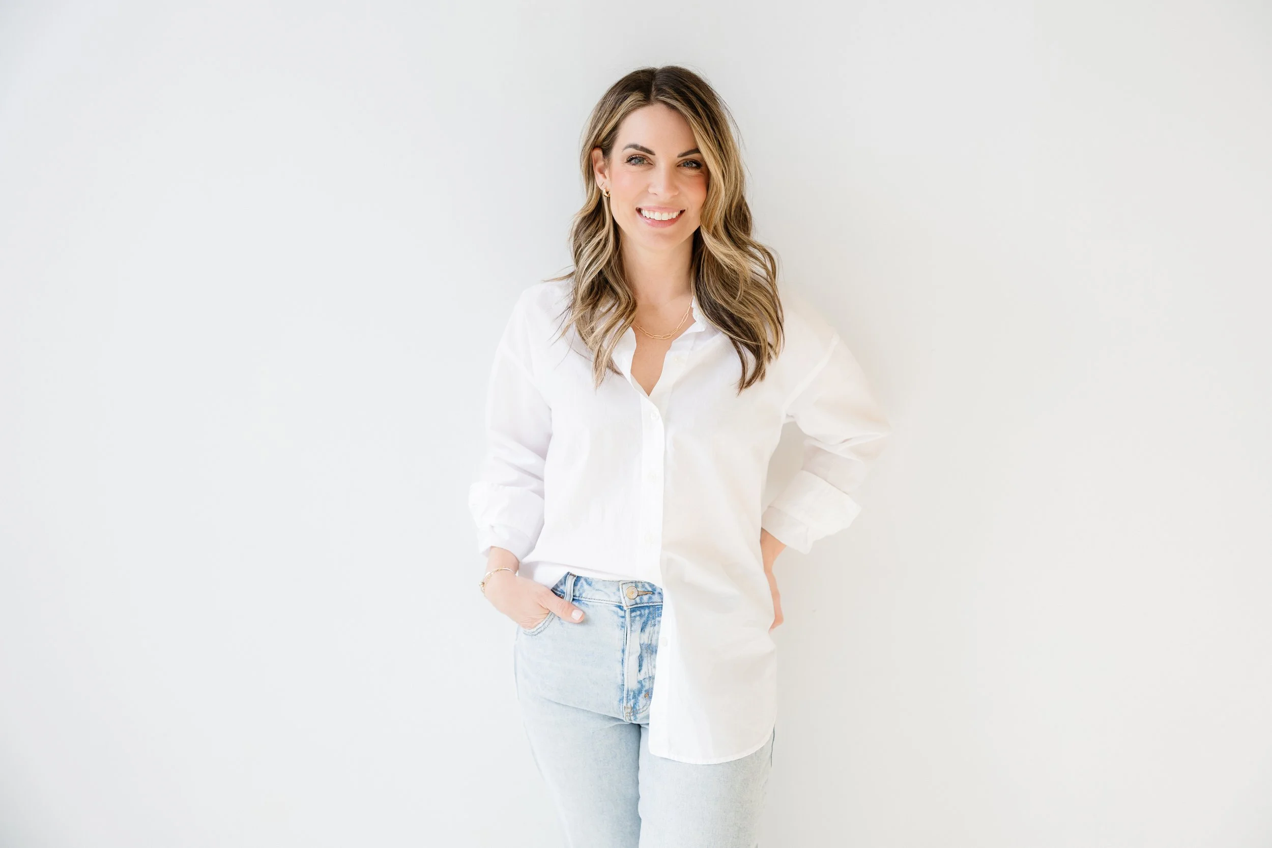 Professional headshot of a woman with wavy light brown hair, wearing a white shirt and light blue jeans, standing in a bright white Anderson Arts Building studio.