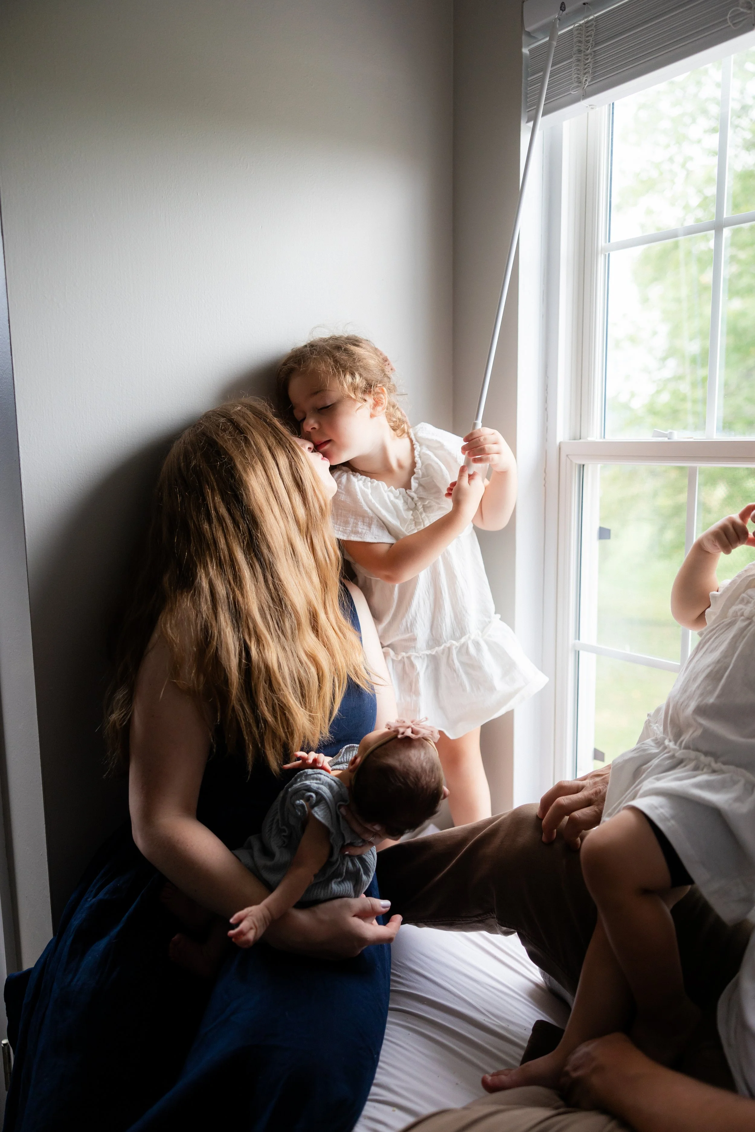 In-home lifestyle newborn photo of a mother holding her baby while siblings share a sweet moment nearby during a family session in Rochester, NY.