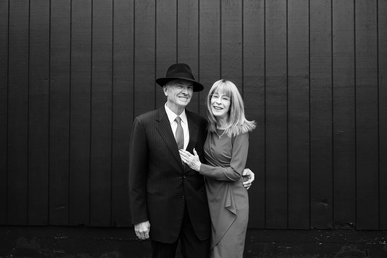 A smiling elderly man in a pinstripe suit and fedora hat stands next to a smiling woman in a dress, both embracing each other, standing against a dark wooden wall.
