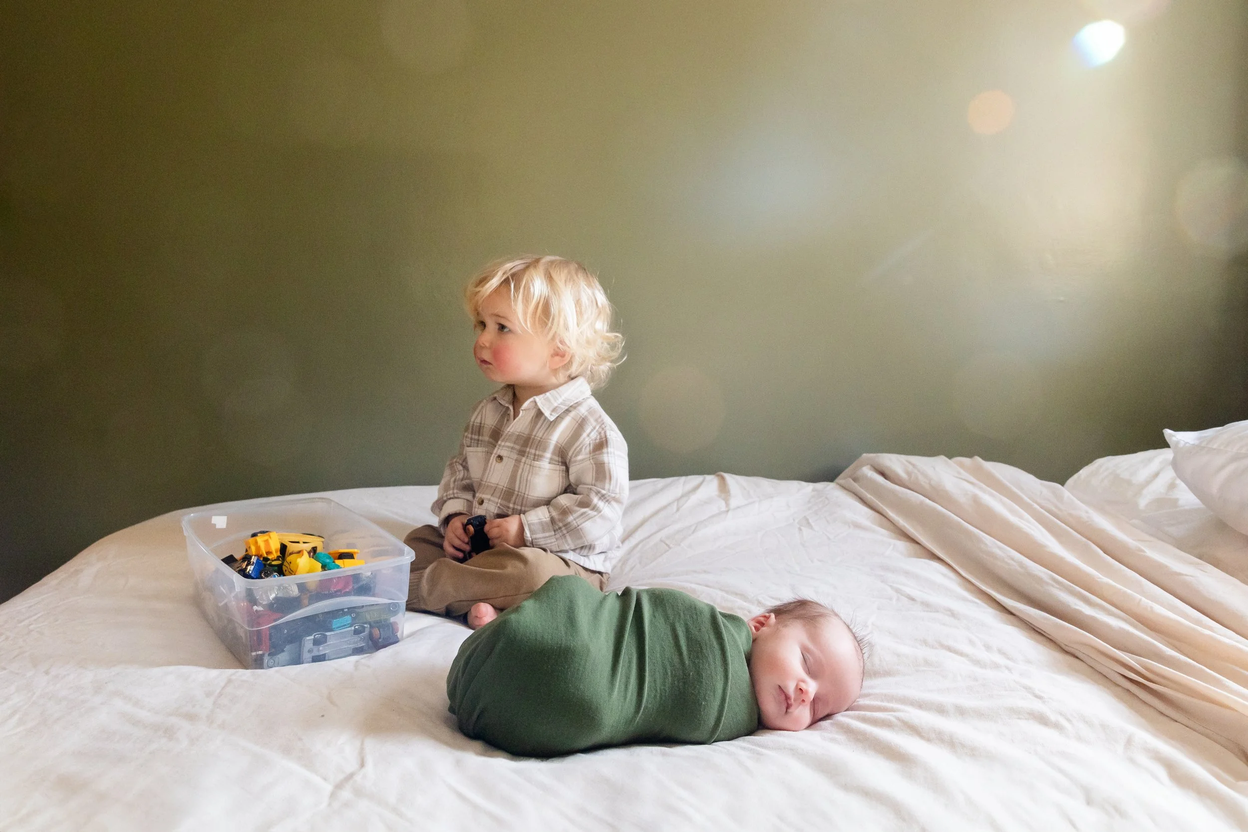 Two young children, one sitting awake playing with toy cars and the other sleeping during an in-home newborn session in Rochester, NY. 