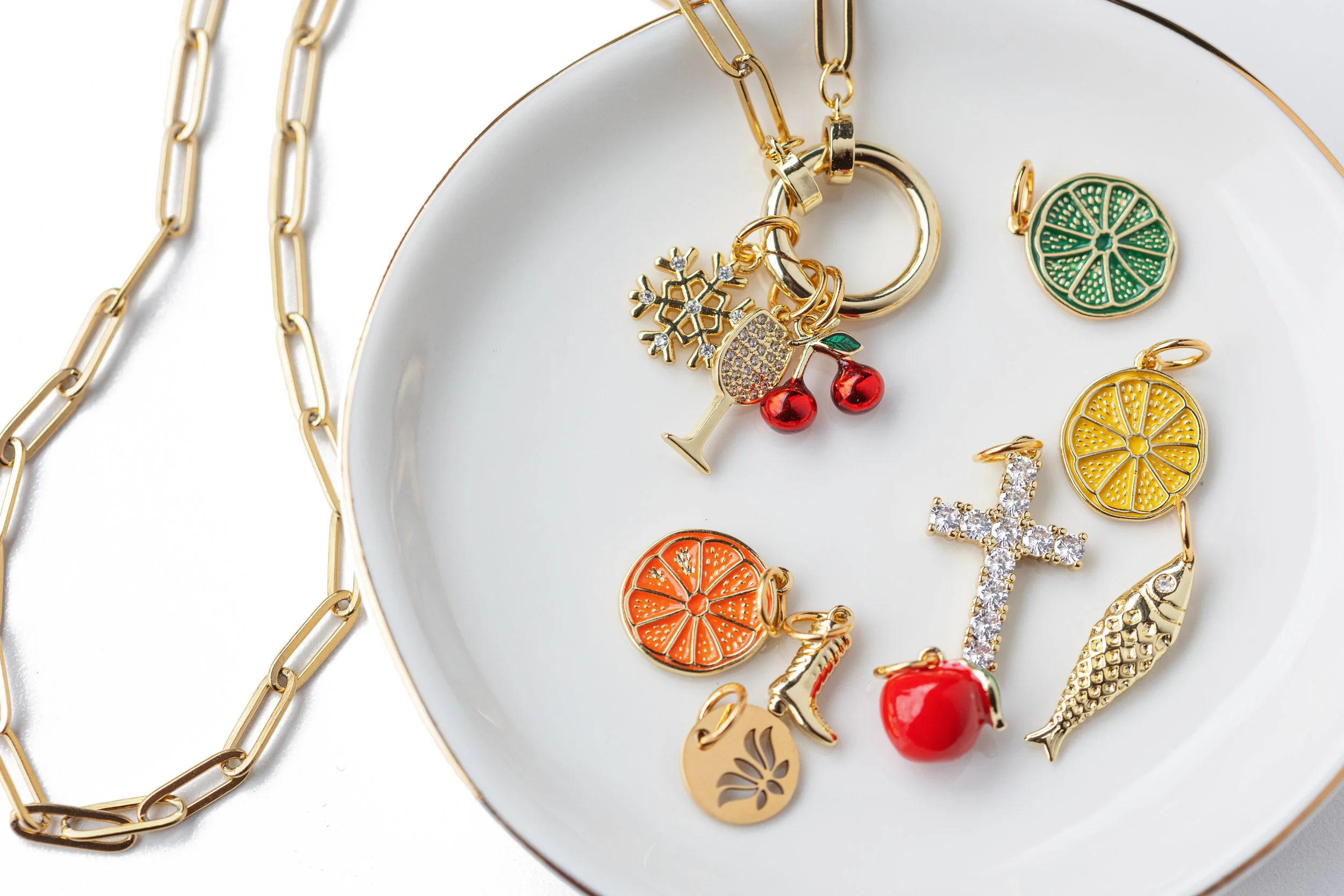 Product photo for Emerson & Oliver featuring gold jewelry charms and a chain necklace styled with fruit and symbolic designs on a neutral surface.
