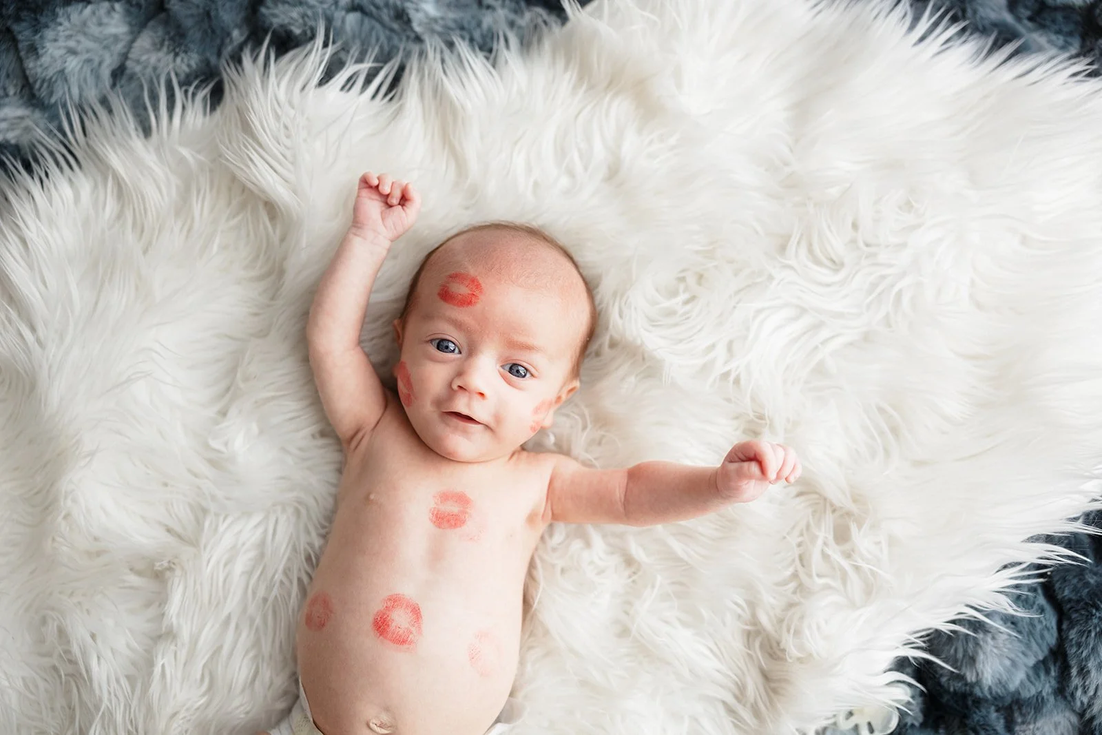 Baby lying on a fluffy white rug with lipstick kiss marks all over his face and body, looking at the camera with arms raised during an in-home newborn session in Rochester, NY.