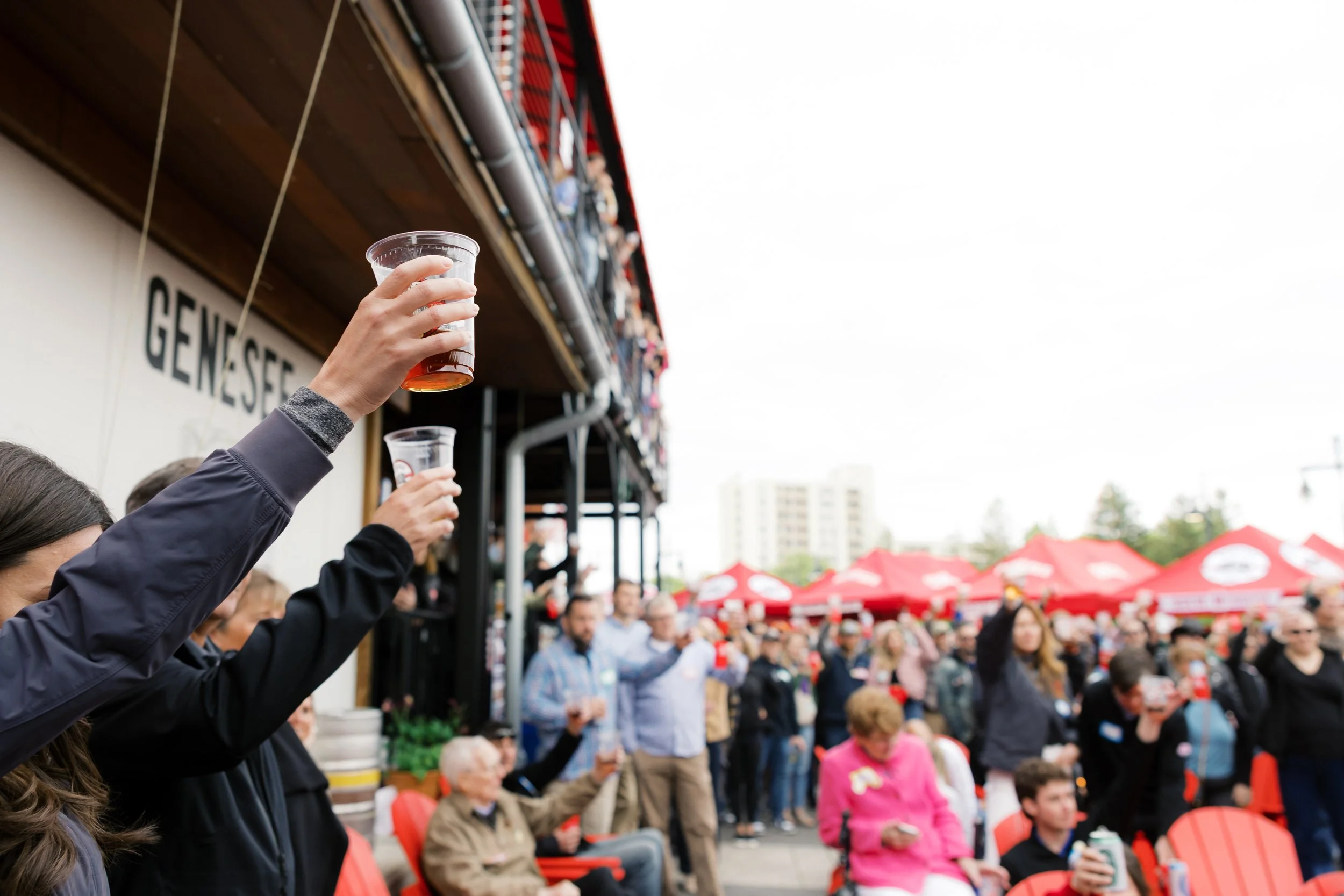 People attending an outdoor event, some holding drinks, sitting and standing under red tents with a crowded background at the Genesee Brew House.