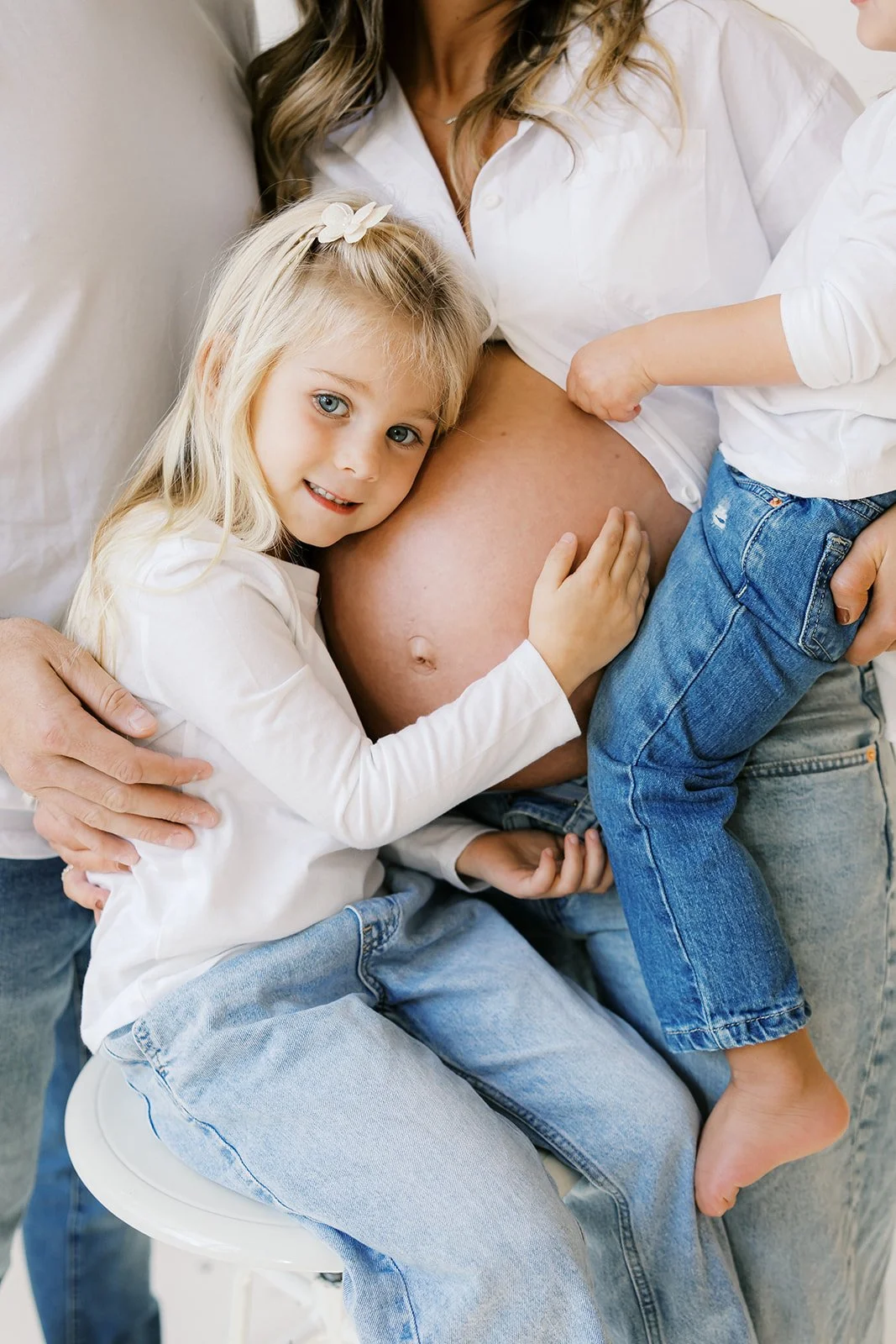 A pregnant woman sitting on a chair, with two young children hugging her belly at a studio session in Rochester, NY.