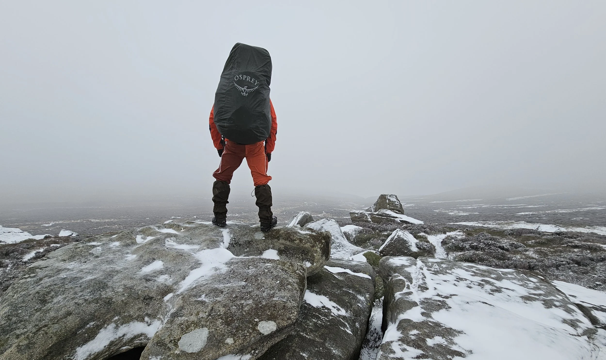 A person stands on rocks in a snowy, foggy landscape, carrying a large black backpack, facing away from the camera.