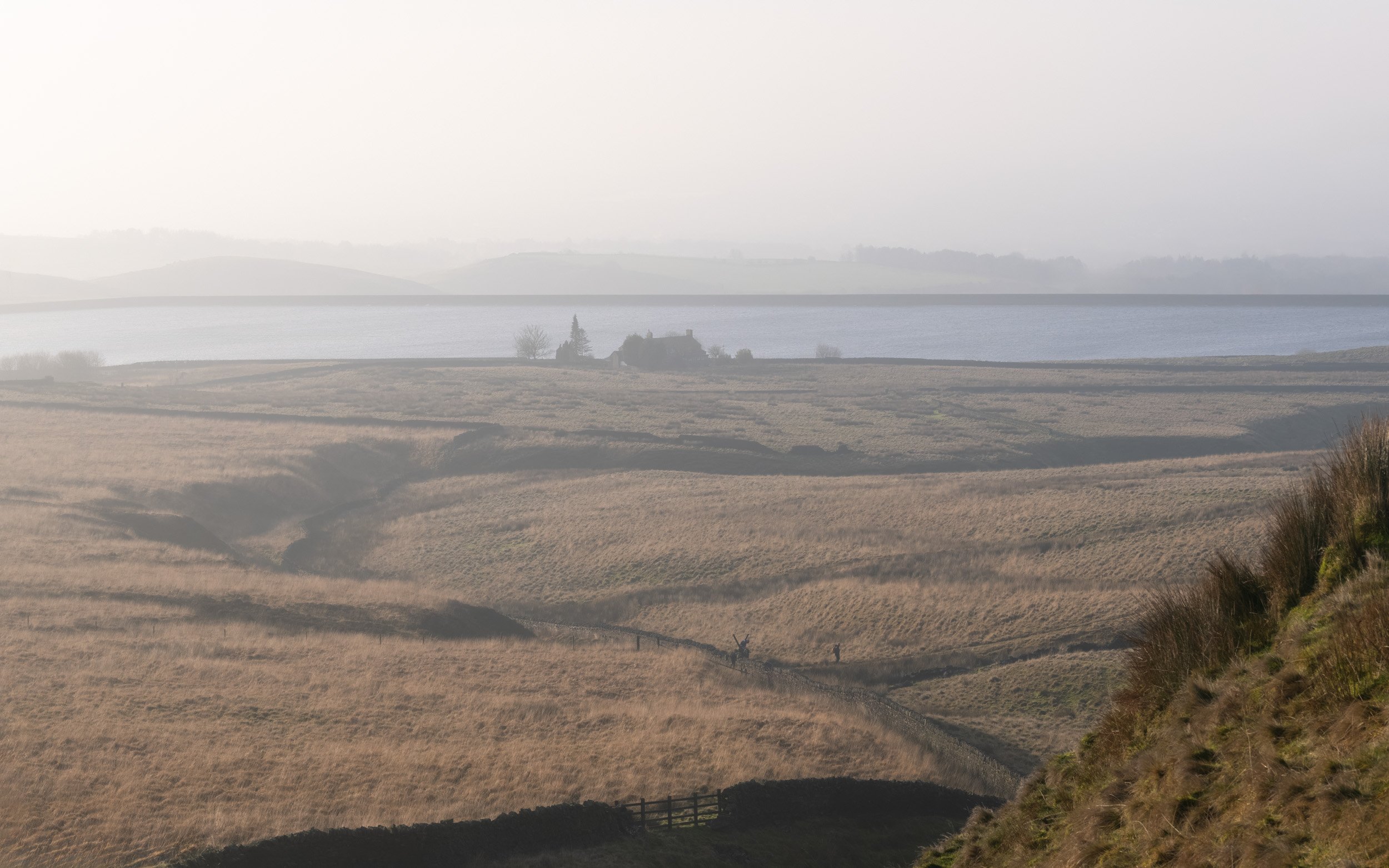 Elevated view across the dry grass slopes of Ashworth Moor towards the Owd Betts pub and Ashworth Moor Reservoir in hazy morning light, with two walkers on the moorland trail.