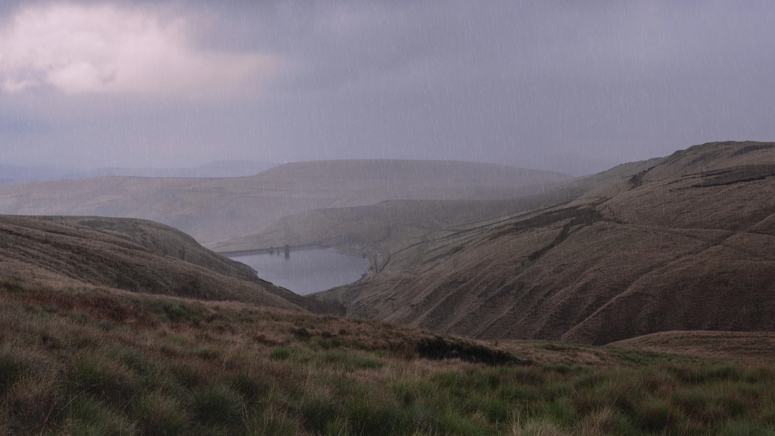 A wide view across rain-streaked moorland hills on Scout Moor looking down into a quiet valley with Scout Moor High Level Reservoir under overcast, atmospheric dark skies.