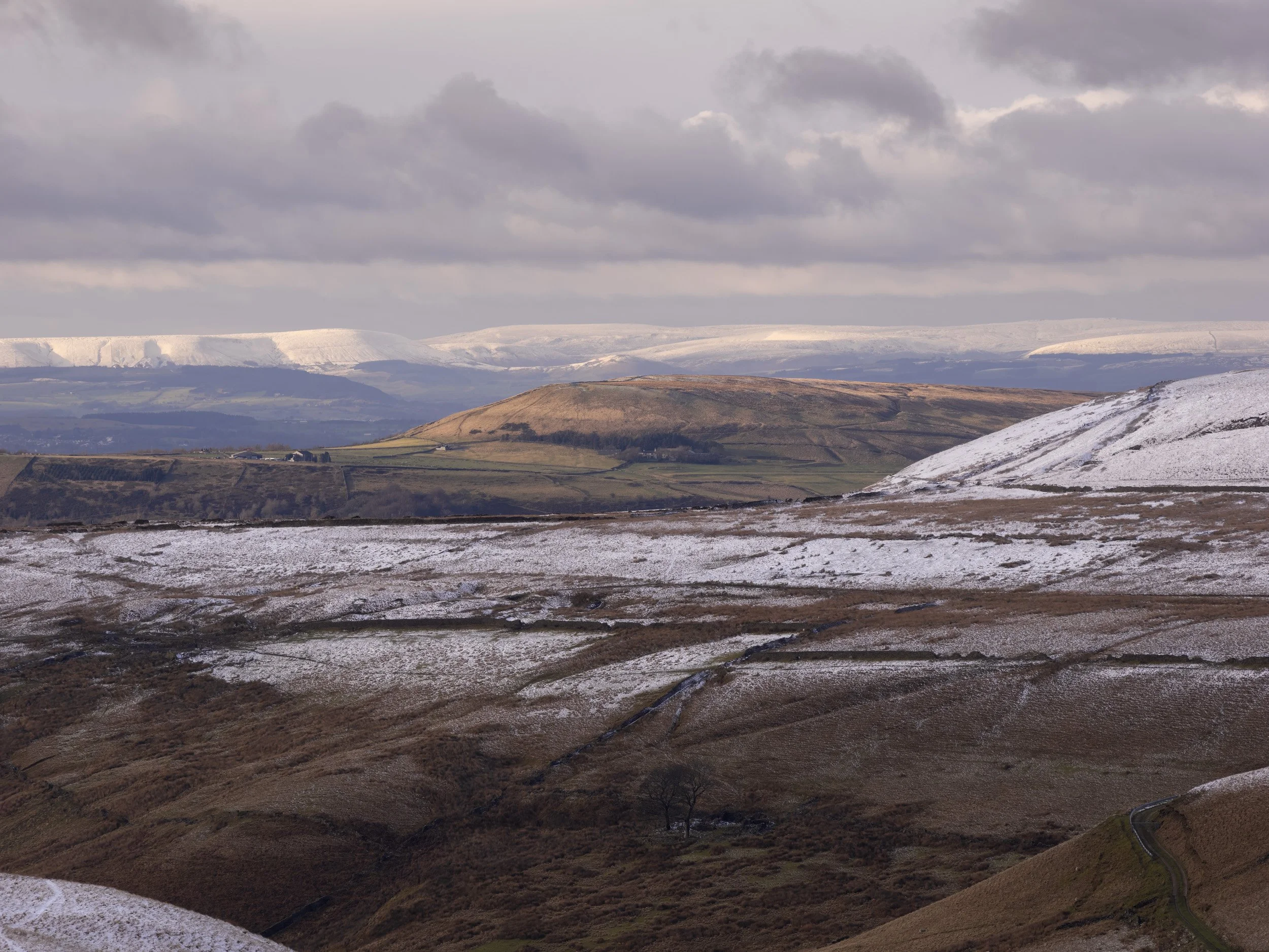 Telephoto vista from above Scout Moor High Level Reservoir showing snow-dusted foreground pastures and sunlit distant snow-capped hills under a cloudy sky.