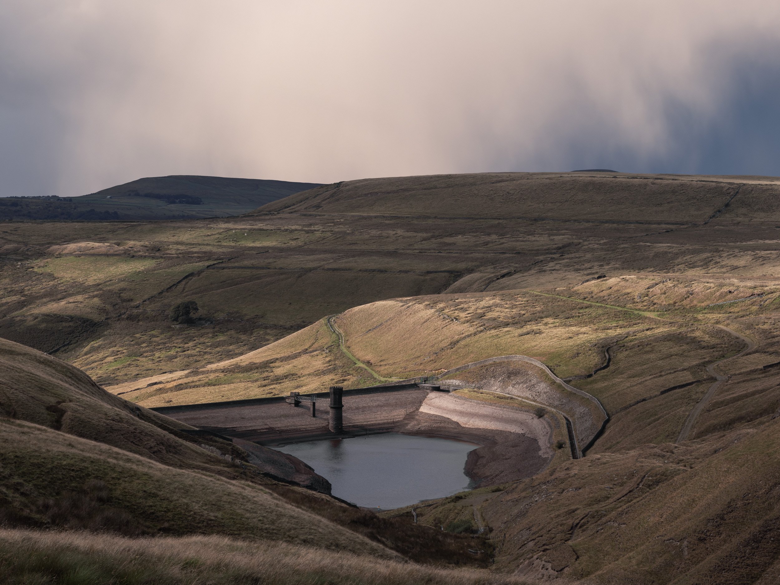 Spotlight of sunshine on the stone reservoir embankment and valley floor under dark grey storm clouds.