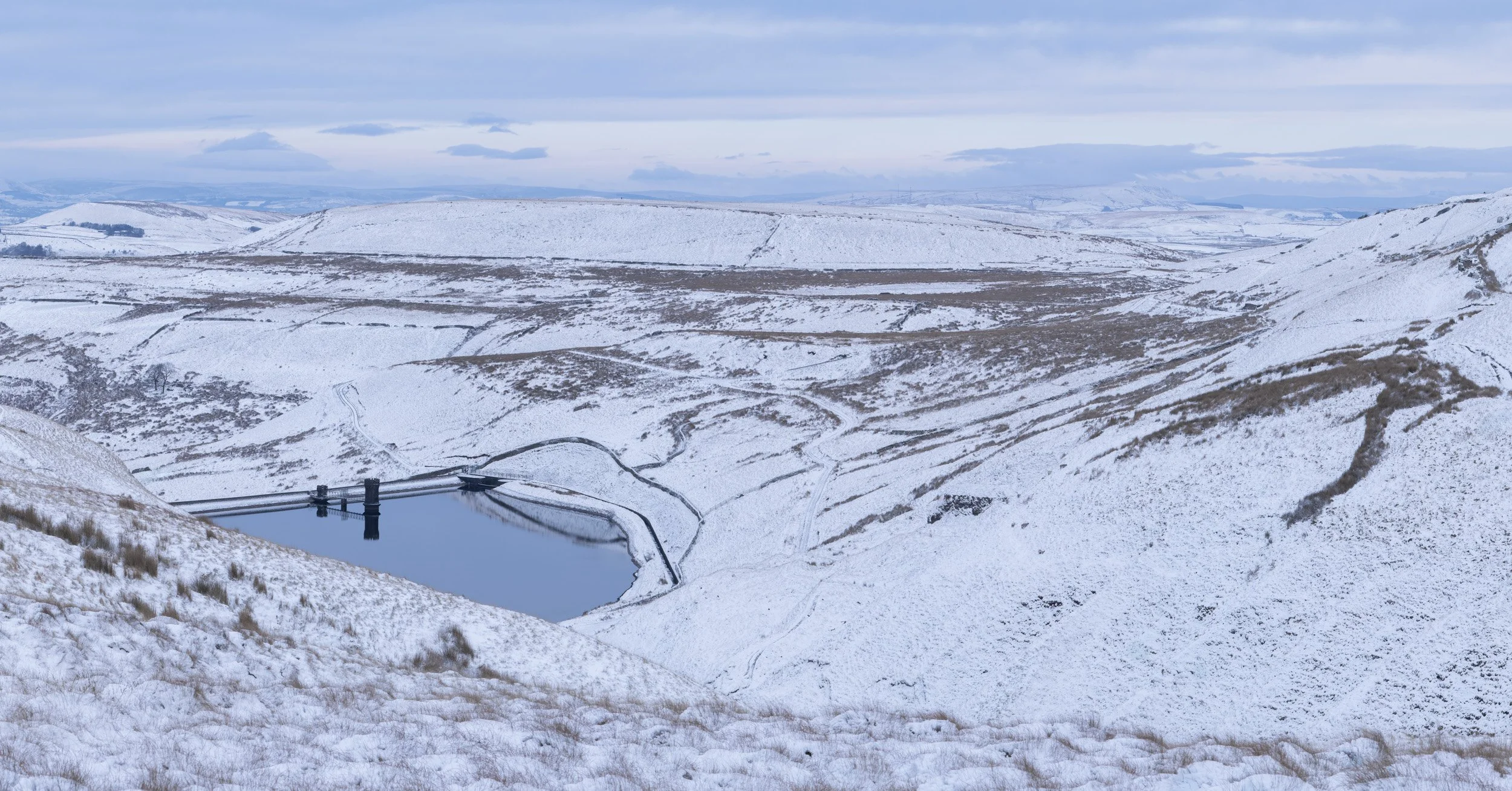 Elevated winter panorama of Scout Moor High Level Reservoir surrounded by snow-covered hills and frosty moorland under a pale sky.