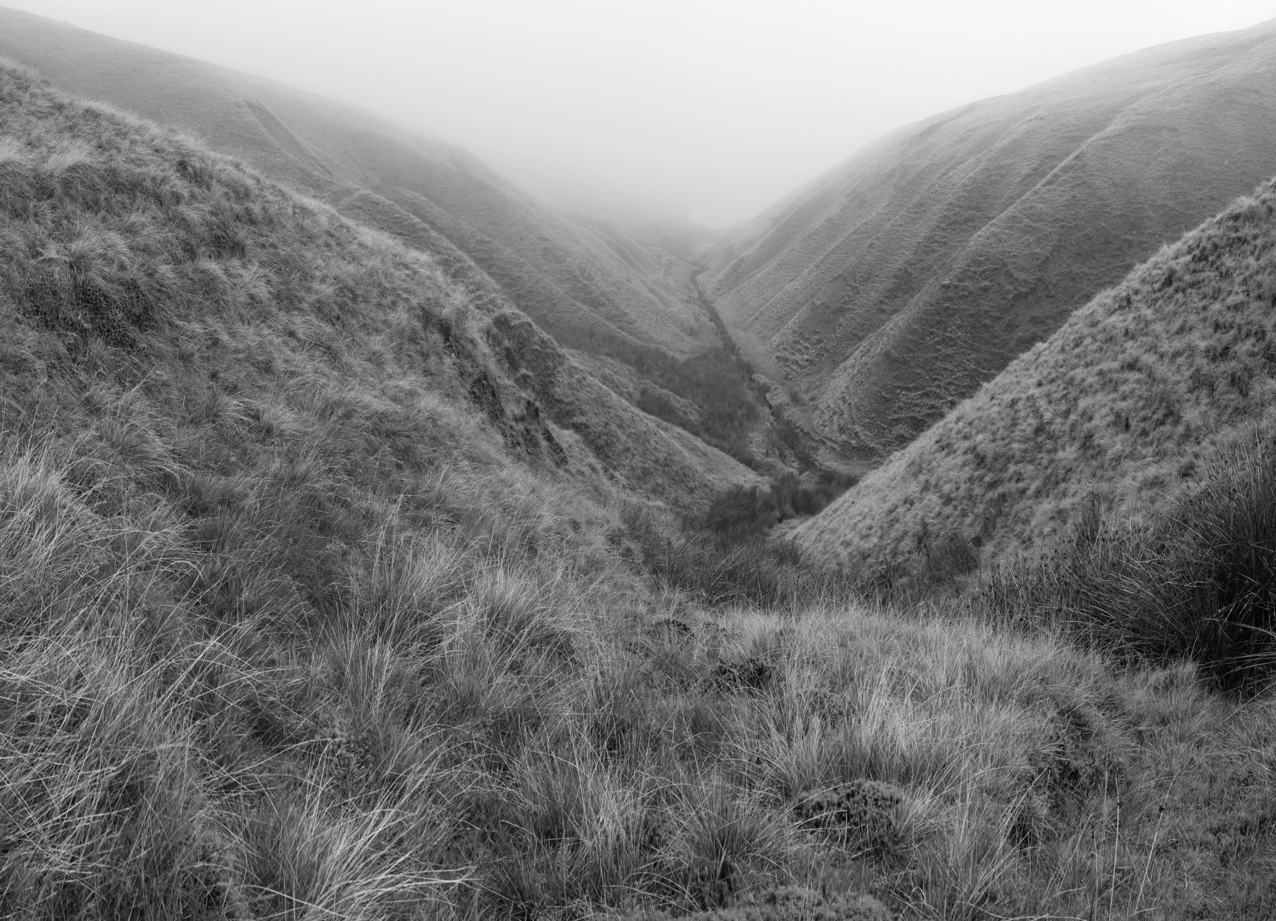 Monochrome high-angle landscape looking down the steep, grassy valley of Scout Moor Brook toward Scout Moor High Level Reservoir.