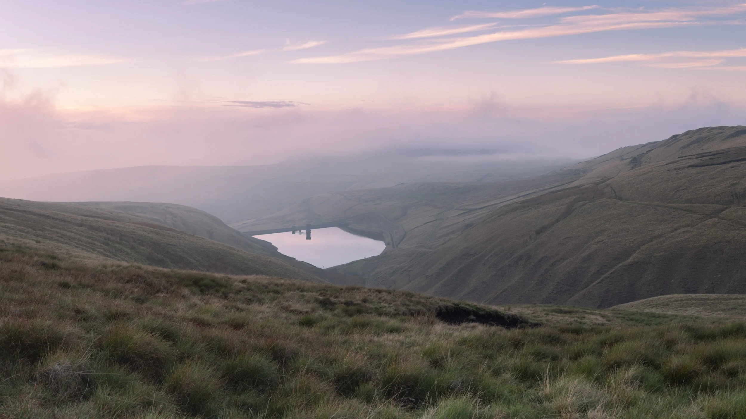 Soft pink twilight sky reflecting in the distant water of Scout Moor High Level Reservoir through light haze.
