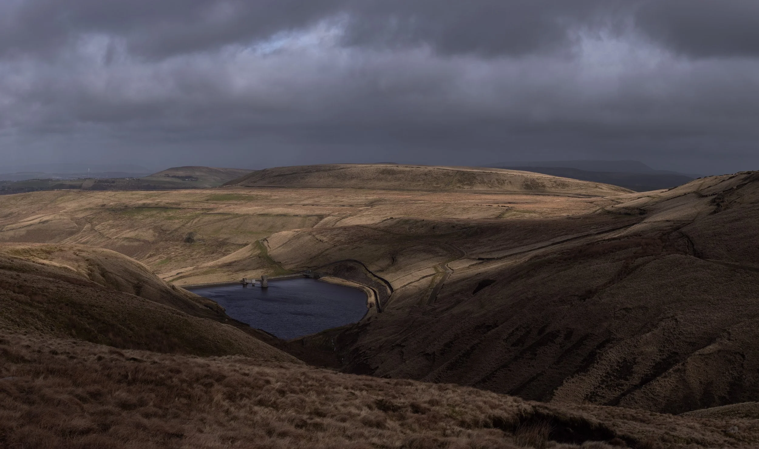 Dark storm clouds looming over the sunlit grass slopes surrounding Scout Moor High Level Reservoir.