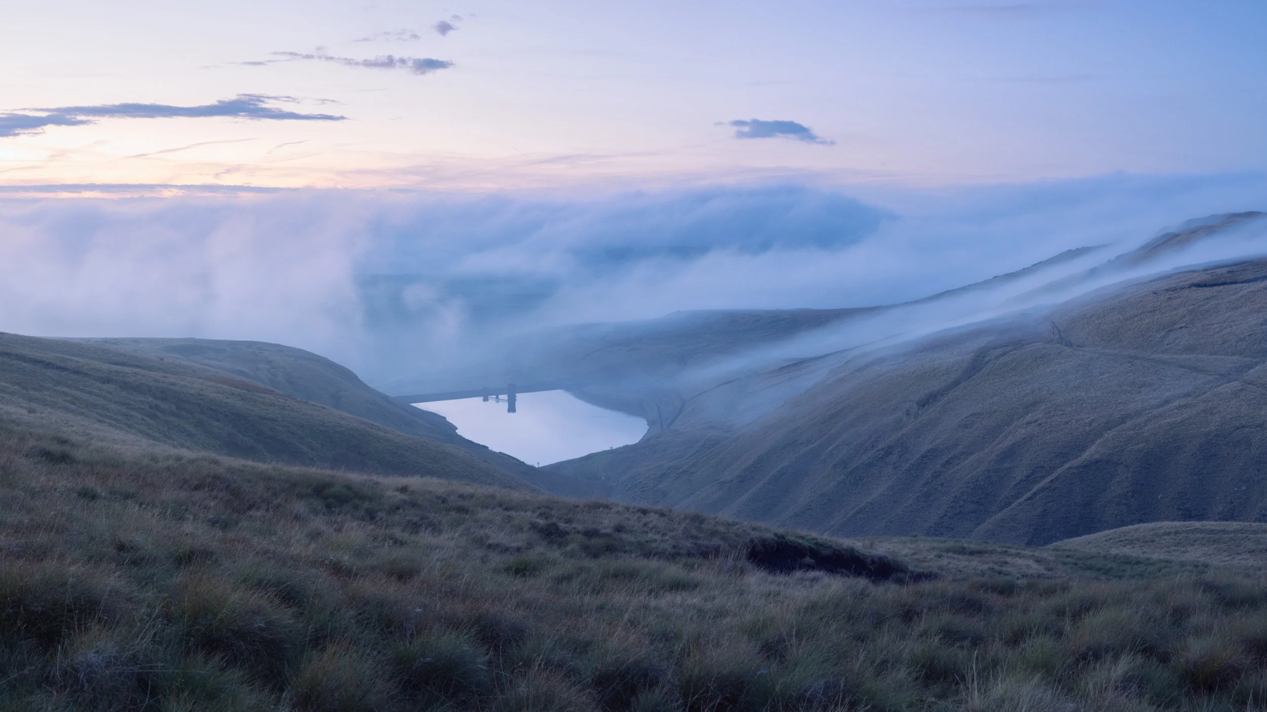 Low fog rolling across the calm surface of Scout Moor High Level Reservoir during twilight.