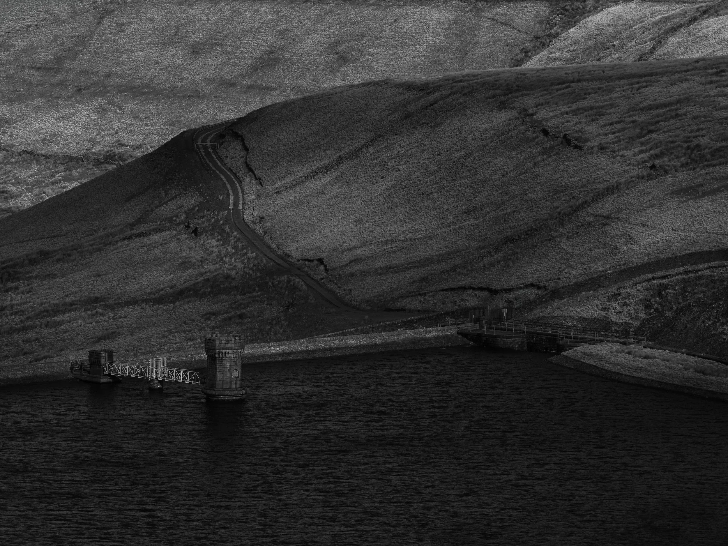 Detailed black and white view of the stone valve tower and footbridge at Scout Moor High Level Reservoir with a curving access track above.