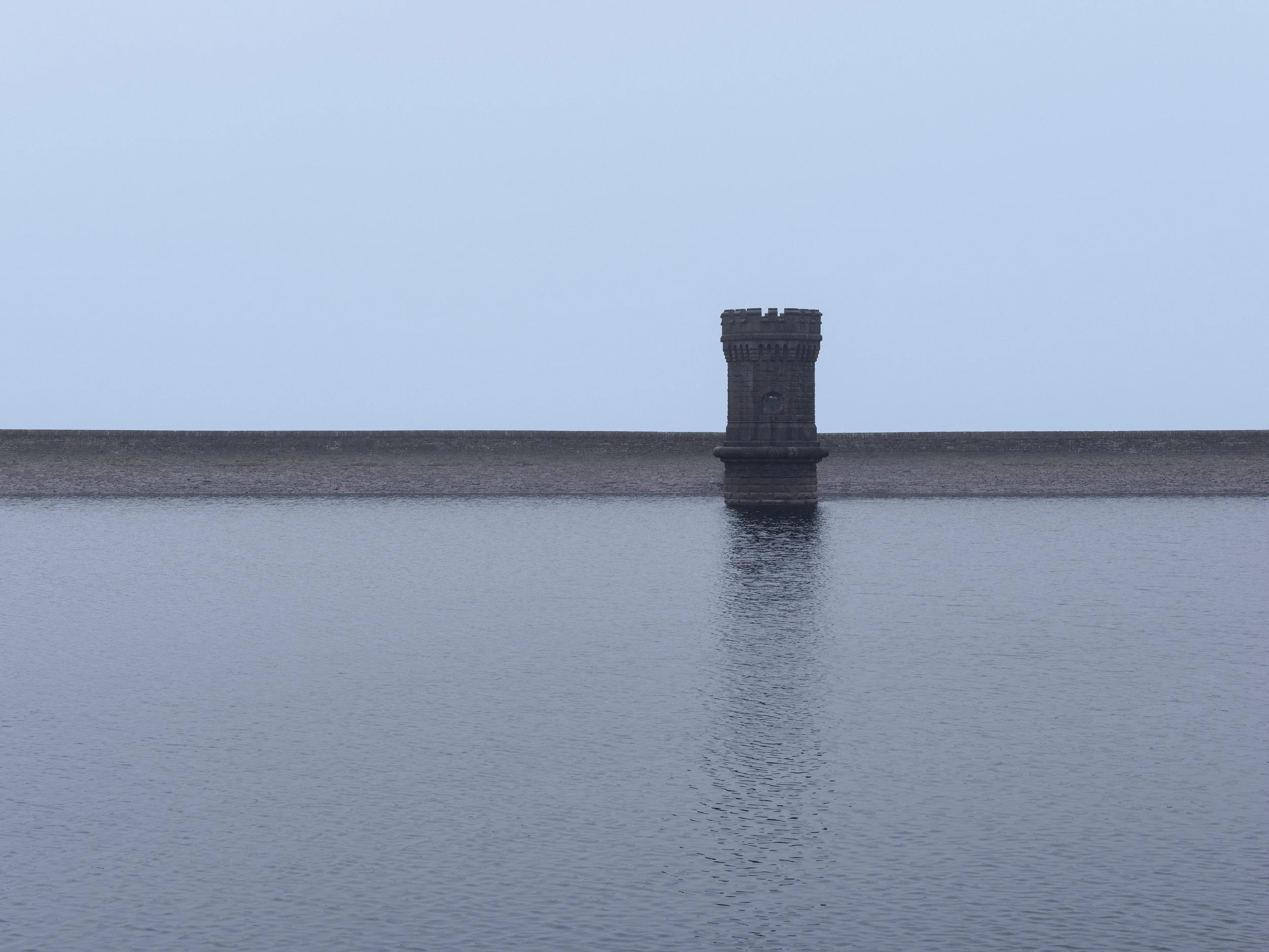 Minimalist composition showing a straight stone embankment wall and Victorian valve tower reflected in still reservoir water.