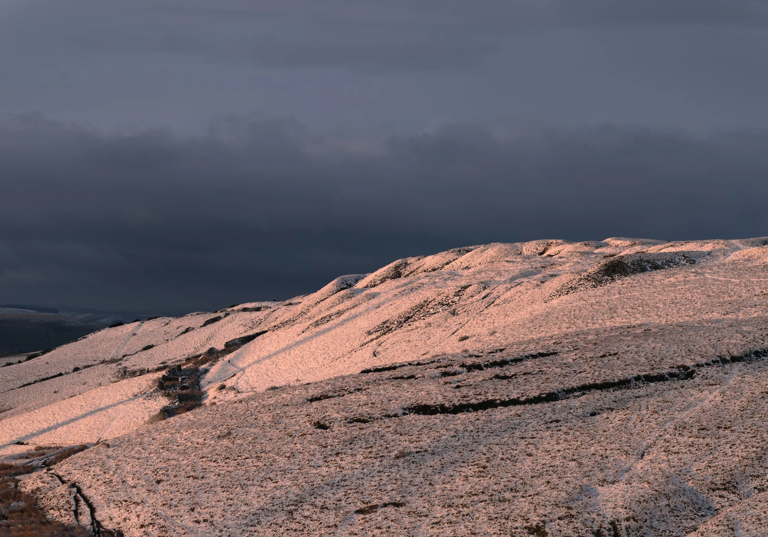 Warm pink dusk light sweeping across the snow-covered slopes of Cragg quarry on Scout Moor against a dark grey winter sky.