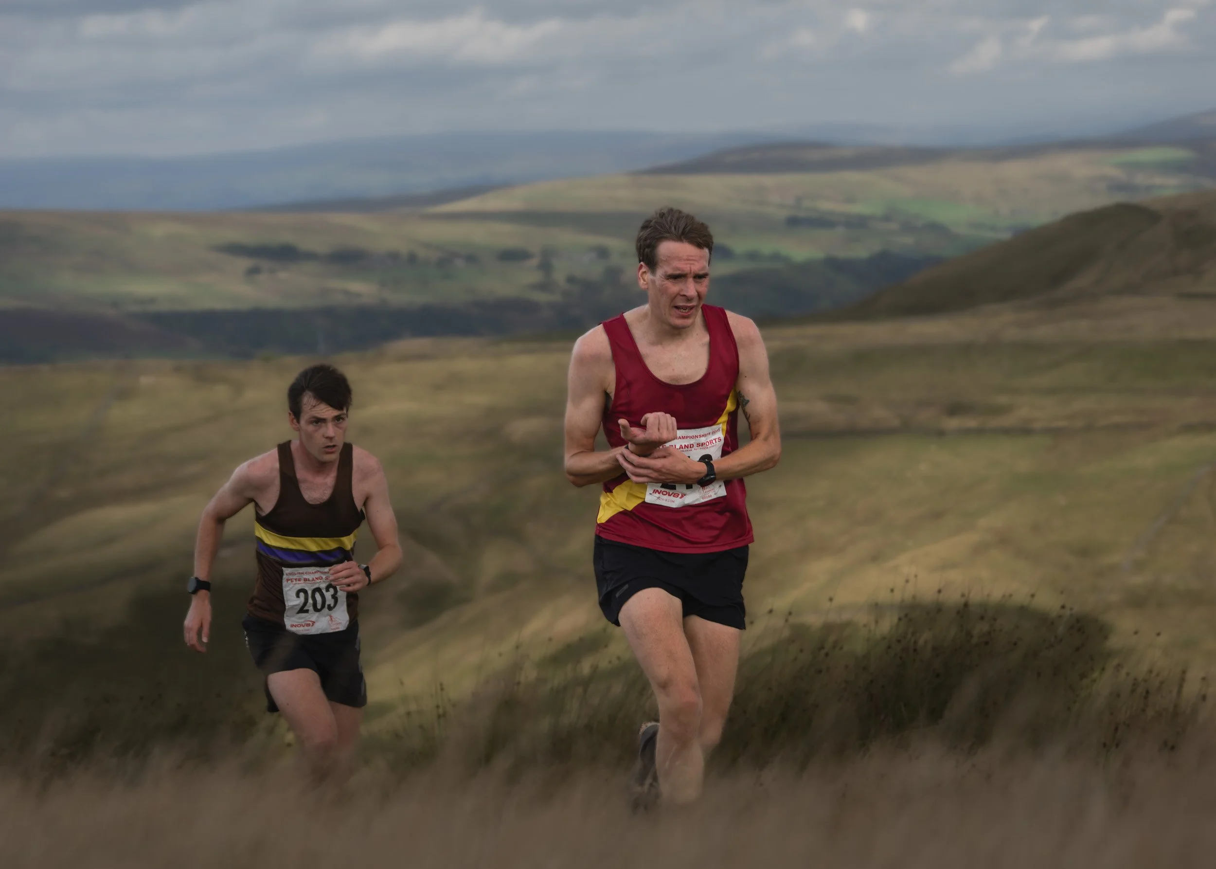 Two fell runners in vest tops charging uphill through moorland grass during the Whittle Pike Fell Race on Scout Moor.