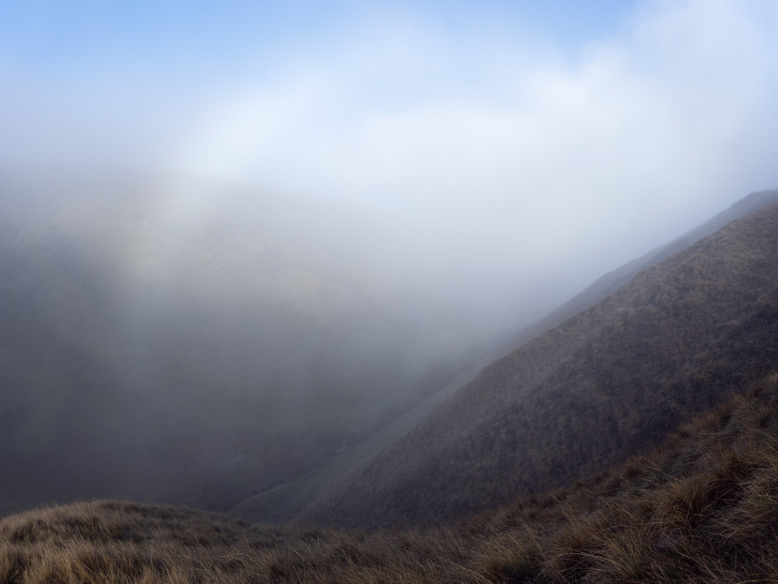 Dramatic landscape photography of thick low-lying mist rolling down steep, grassy moorland slopes in the valley near Scout Moor High Level Reservoir under a soft blue sky.