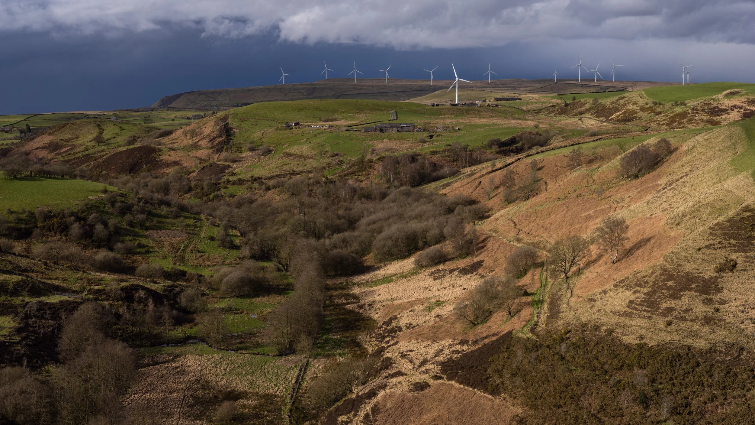 Wide view looking up the wooded slopes of Cheesden Valley from Birtle towards the wind turbines of Scout Moor Wind Farm under a moody storm sky."