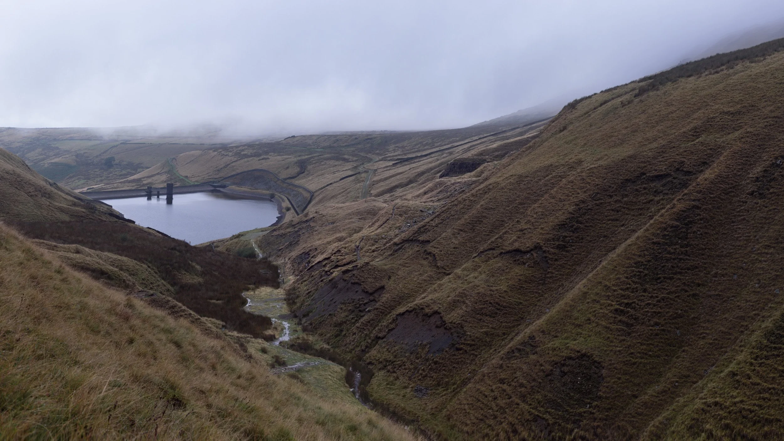 Narrow stream winding down a steep moorland ravine toward Scout Moor High Level Reservoir under mist.
