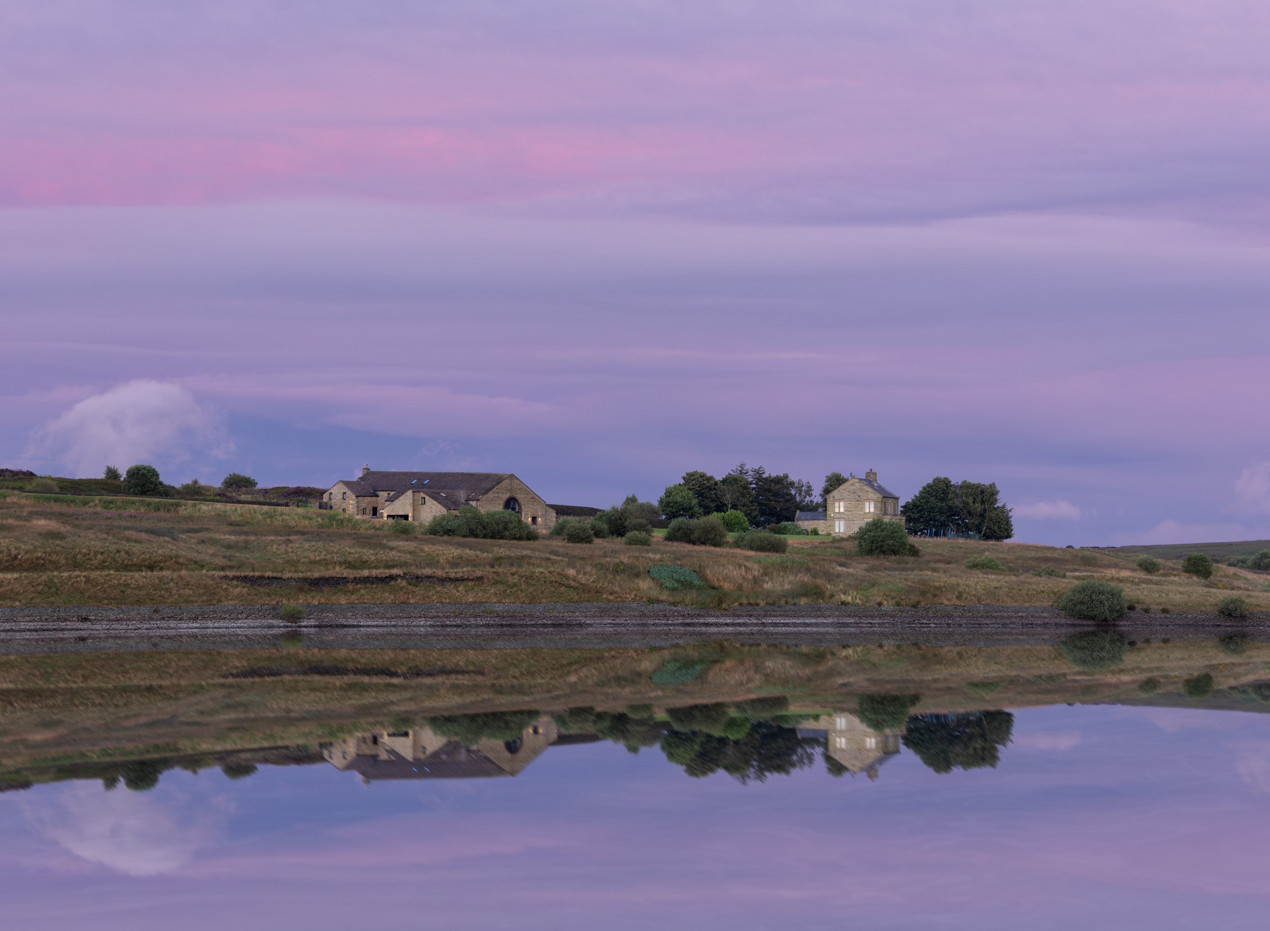 A rural landscape with houses, trees, and a body of water reflecting the sky at dusk with pink and purple clouds.