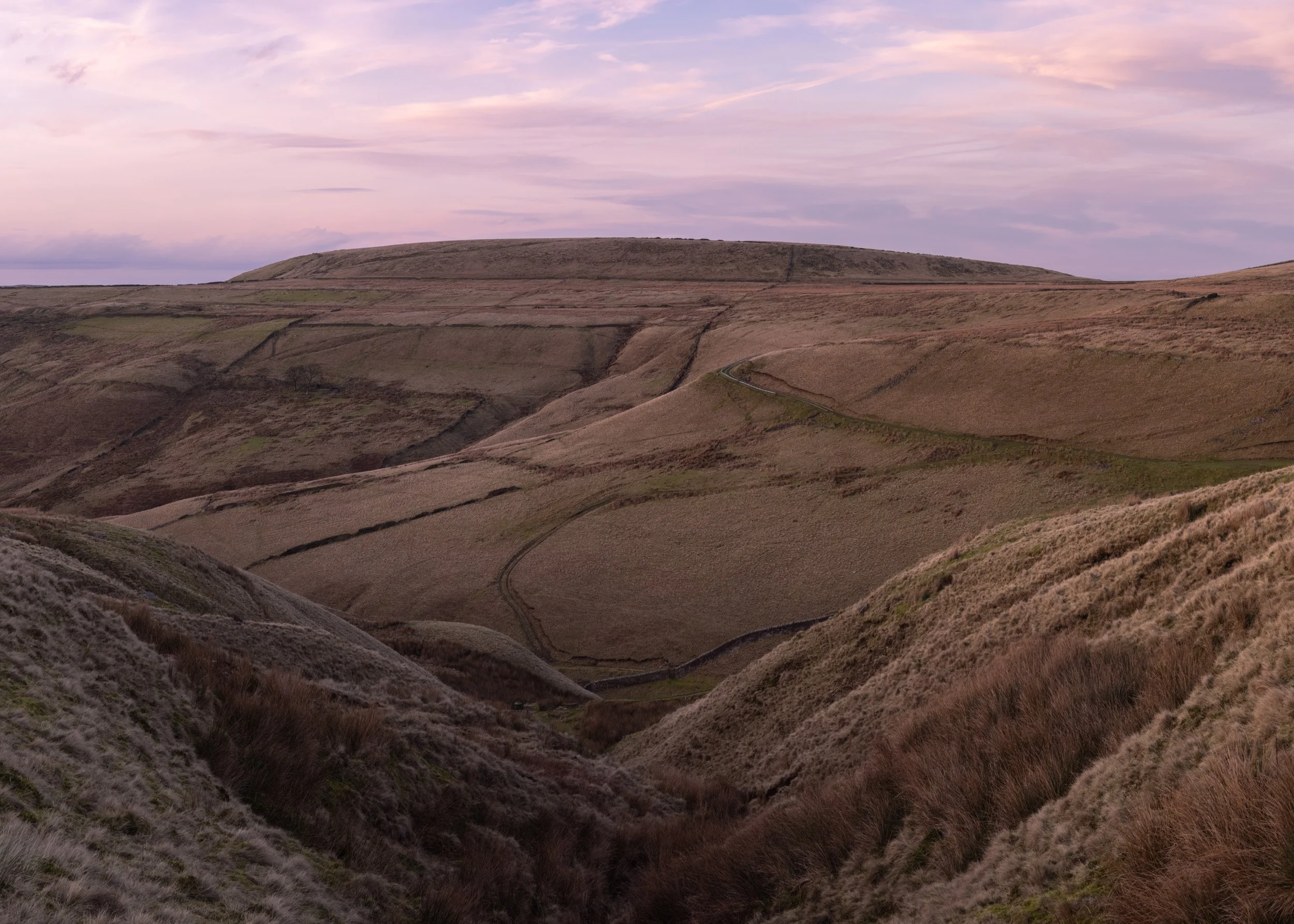 Deep V-shaped valley in the foreground leading across quiet moorland slopes toward the flat-topped ridge of Cowpe Lowe under a soft pink pastel sky on Scout Moor.