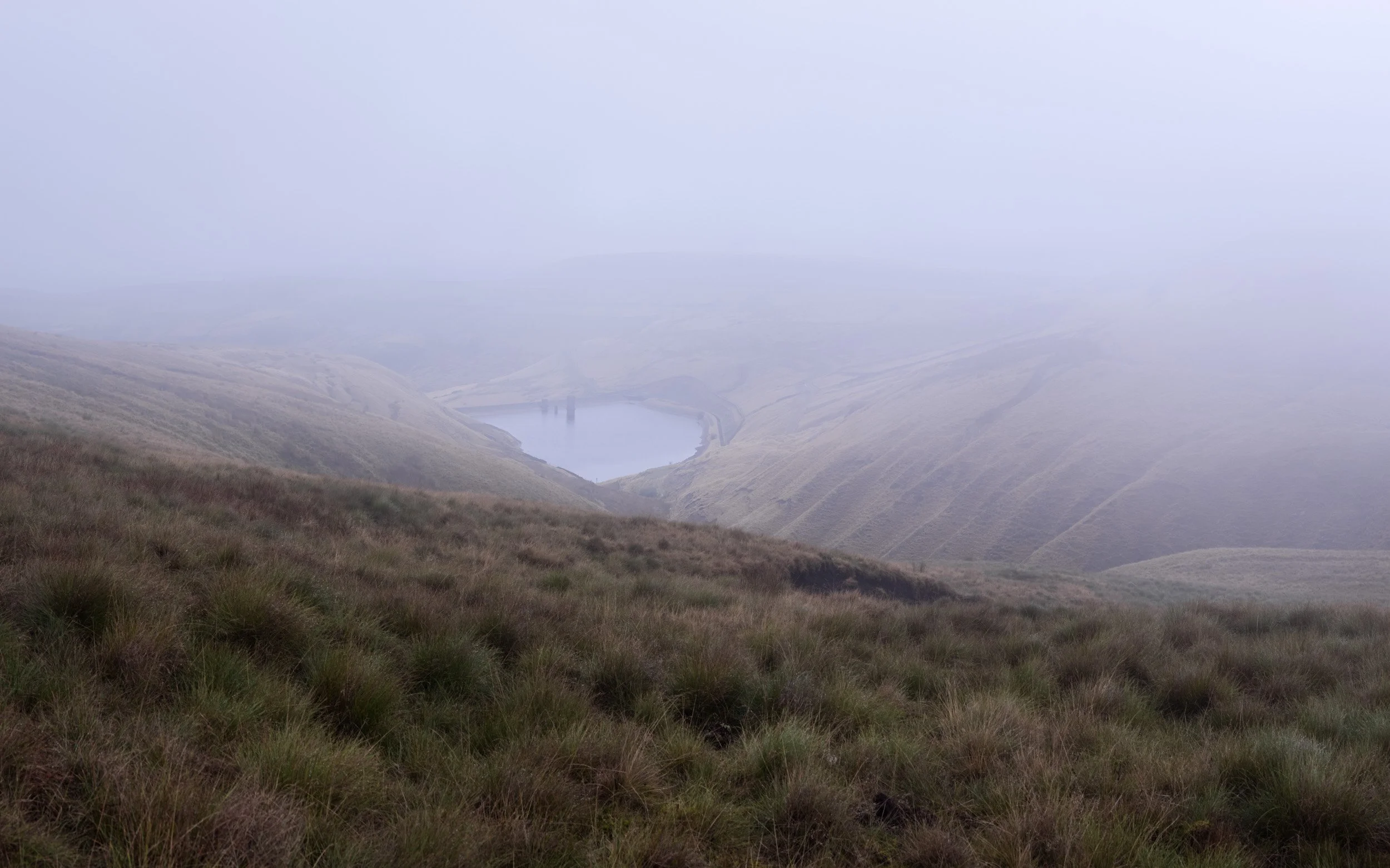 Soft white mist obscuring the distant hills around Scout Moor High Level Reservoir seen from a grassy foreground.