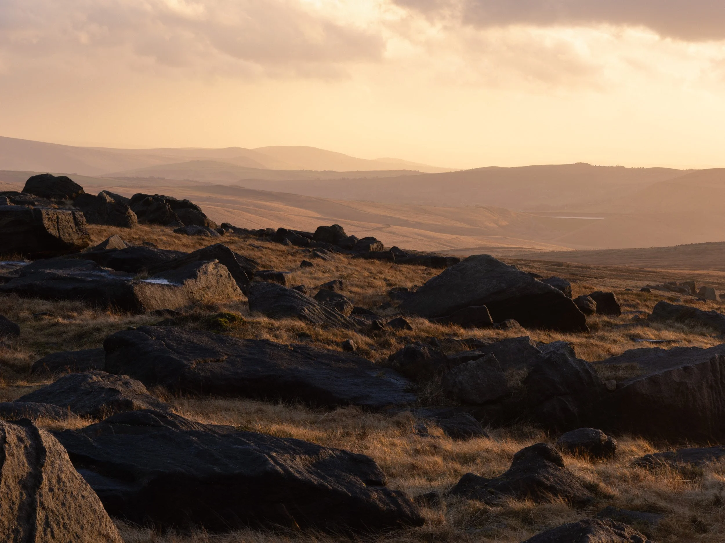 Scattered gritstone boulders on Blackstone Edge near Littleborough, illuminated by golden sunset light looking out toward layered Pennine hills.