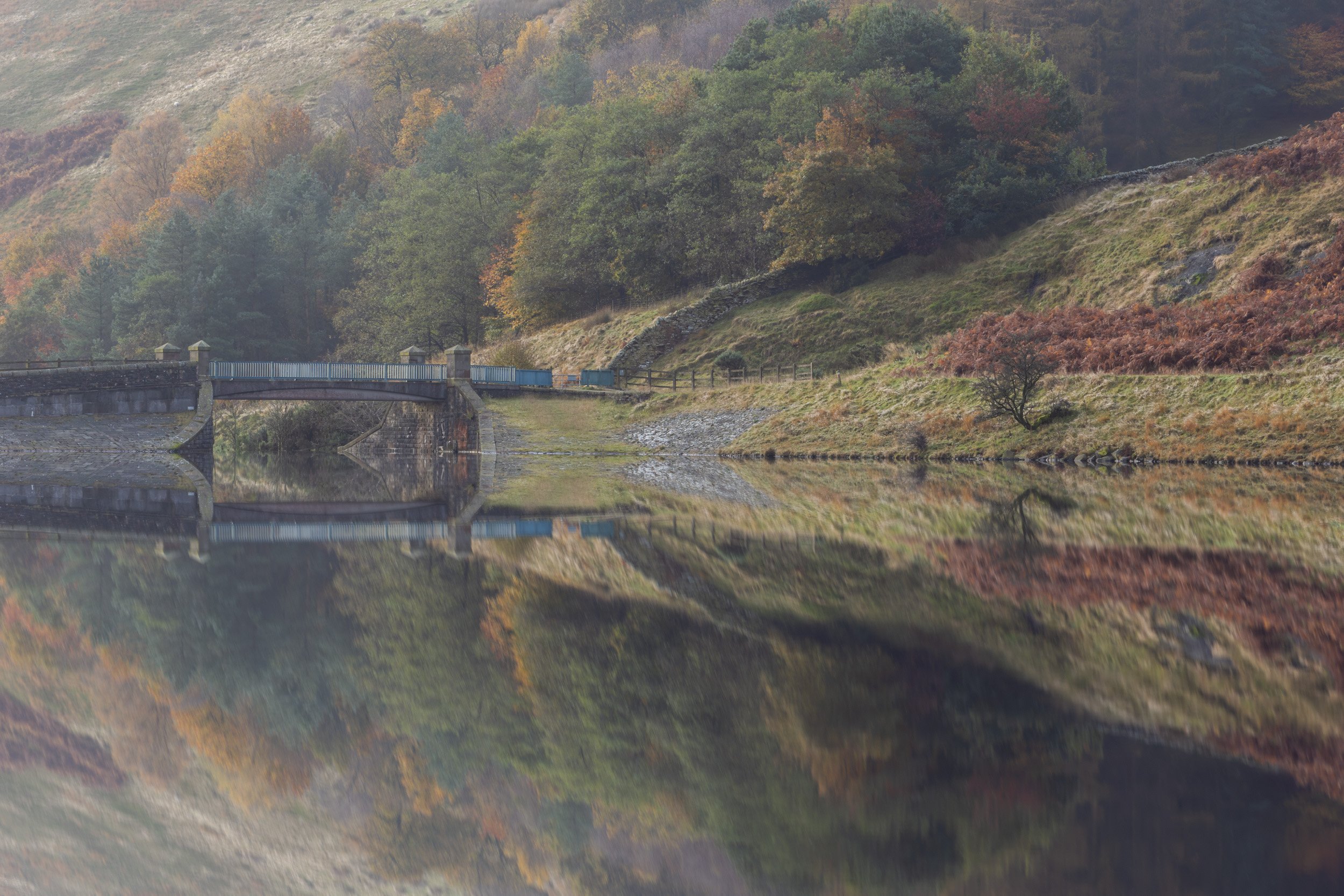 Perfect water reflection of a stone bridge, drystone wall, and autumn-colored hillside at Naden Valley Reservoirs near Scout Moor.