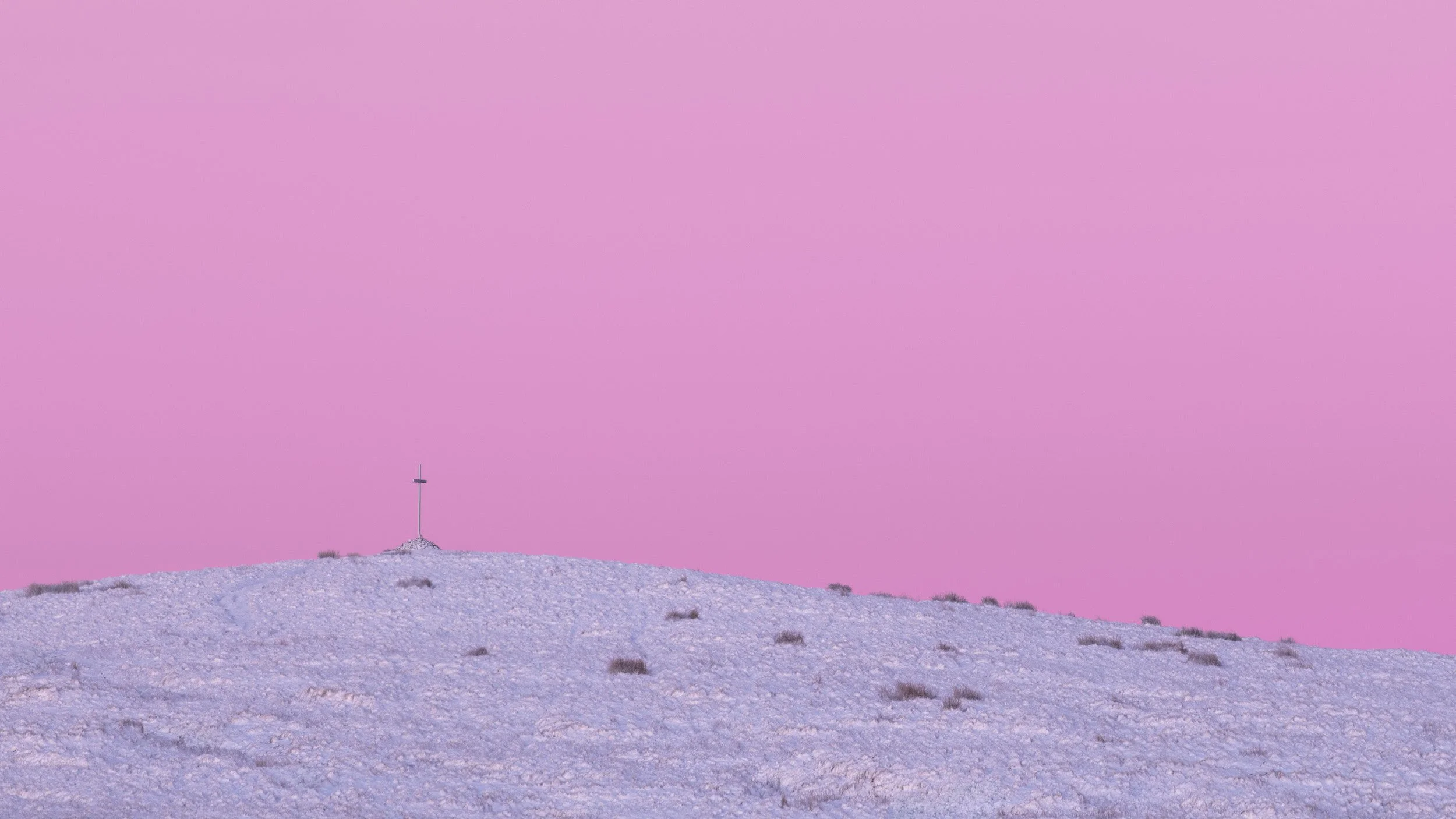 Silhouetted summit cross atop the snow-covered peak of Whittle Pike on Scout Moor against a solid pastel pink sky.