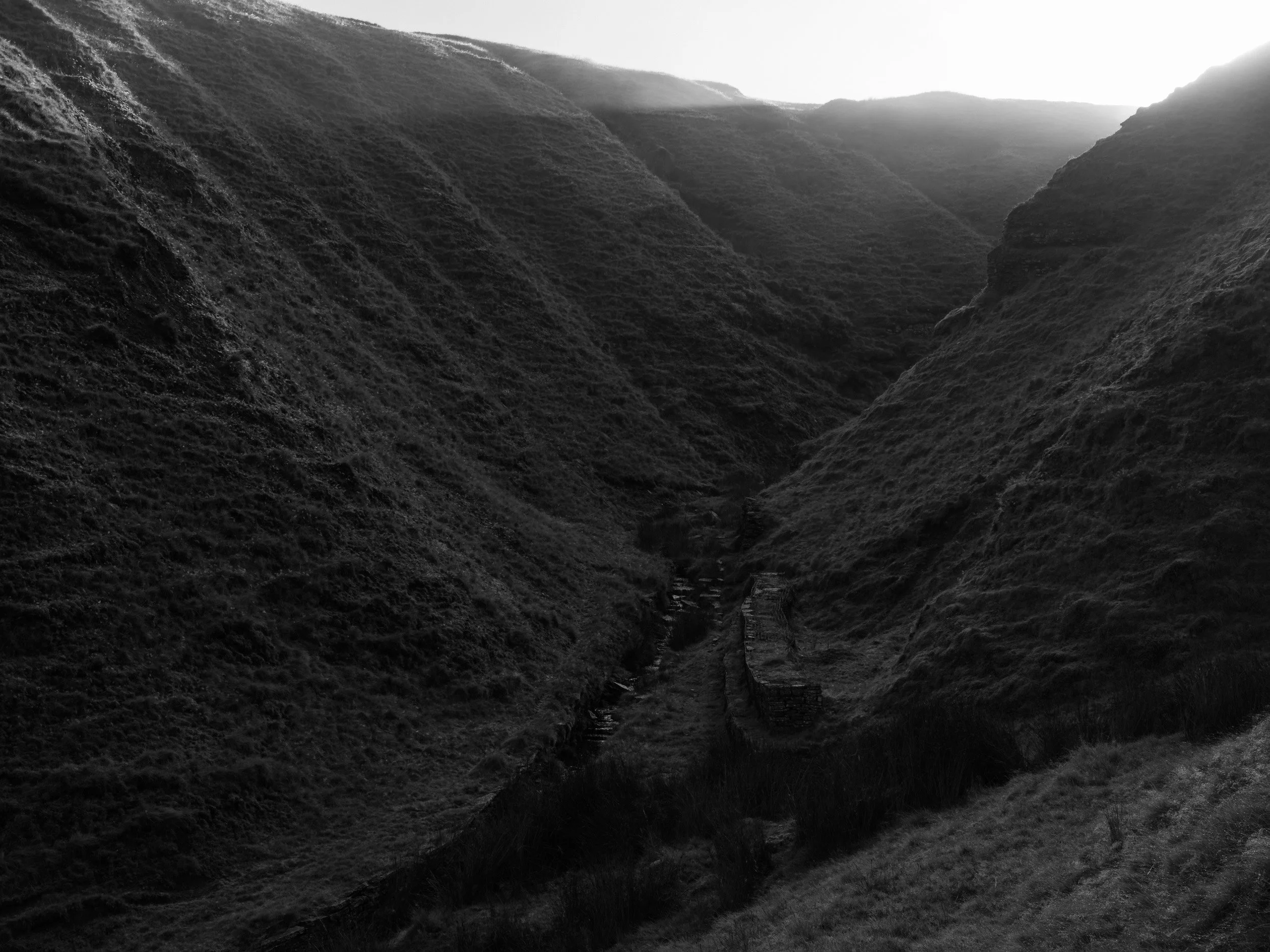 High-contrast monochrome shot looking down into the dark, steep ravine of Scout Moor Brook with light breaking over the ridge.