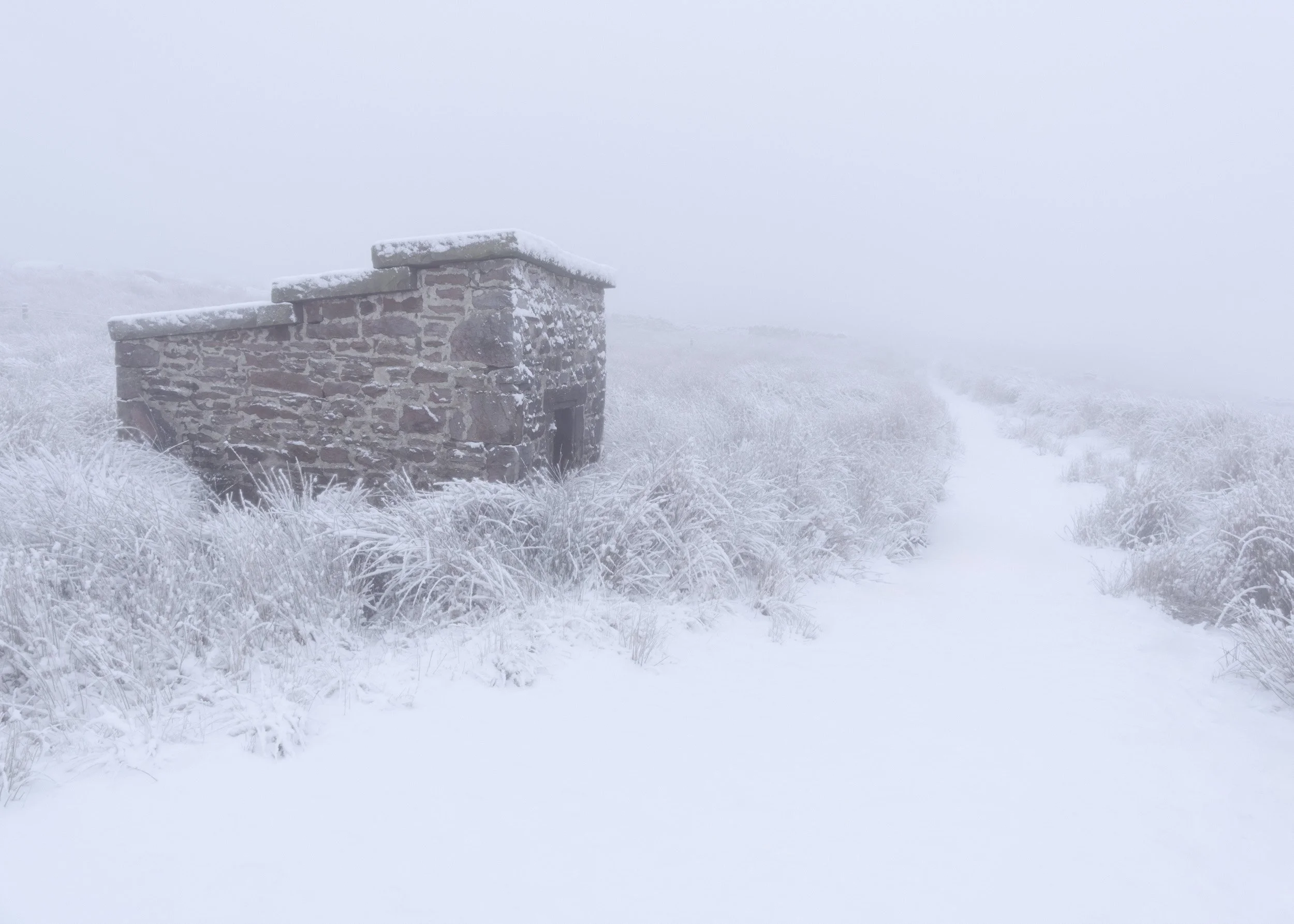 Small stone quarry building standing isolated beside a snow-covered path on a foggy winter day on Scout Moor near Rochdale.
