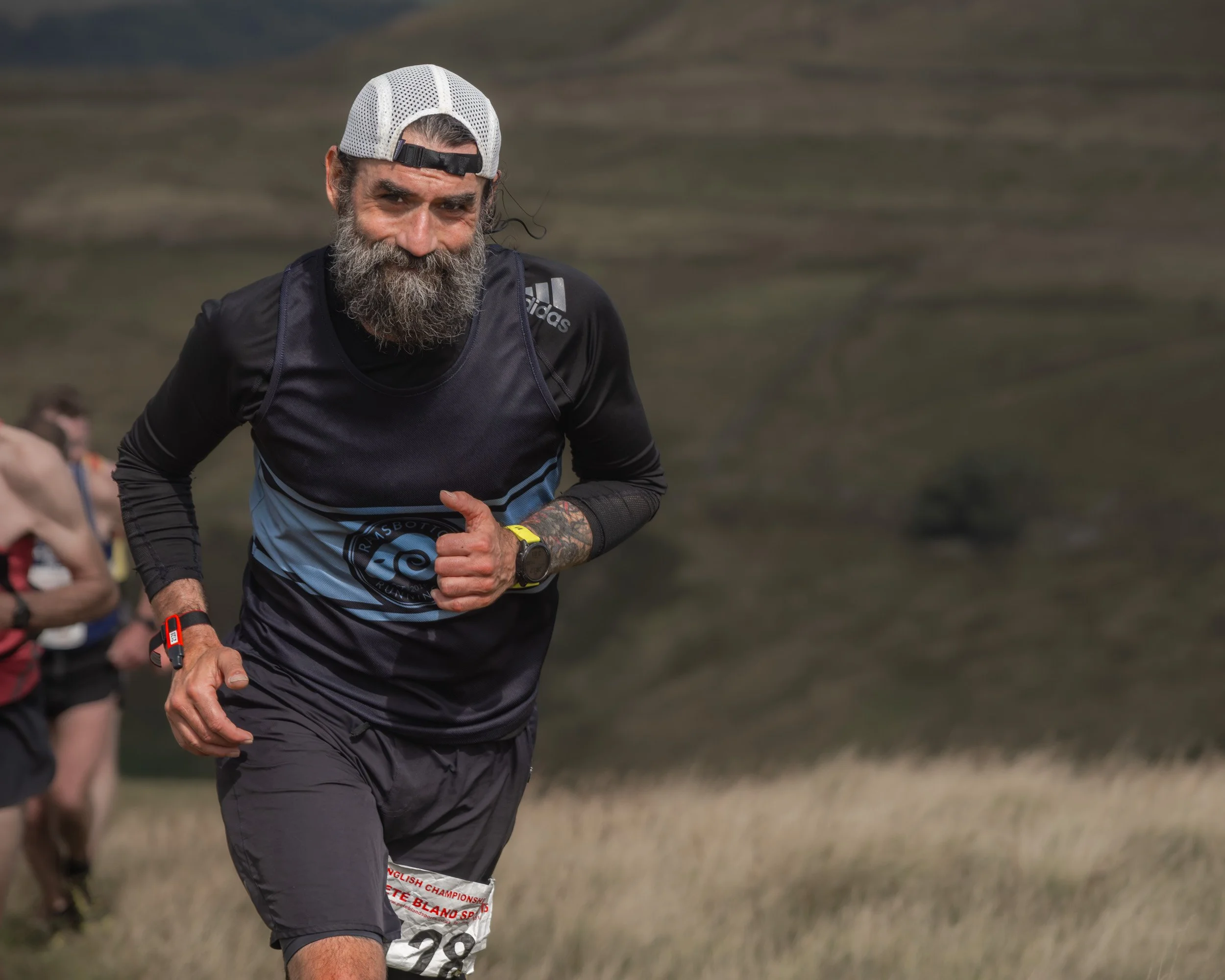 Close-up of a smiling, bearded fell runner in a black long-sleeve top giving a thumbs-up mid-stride on Scout Moor.