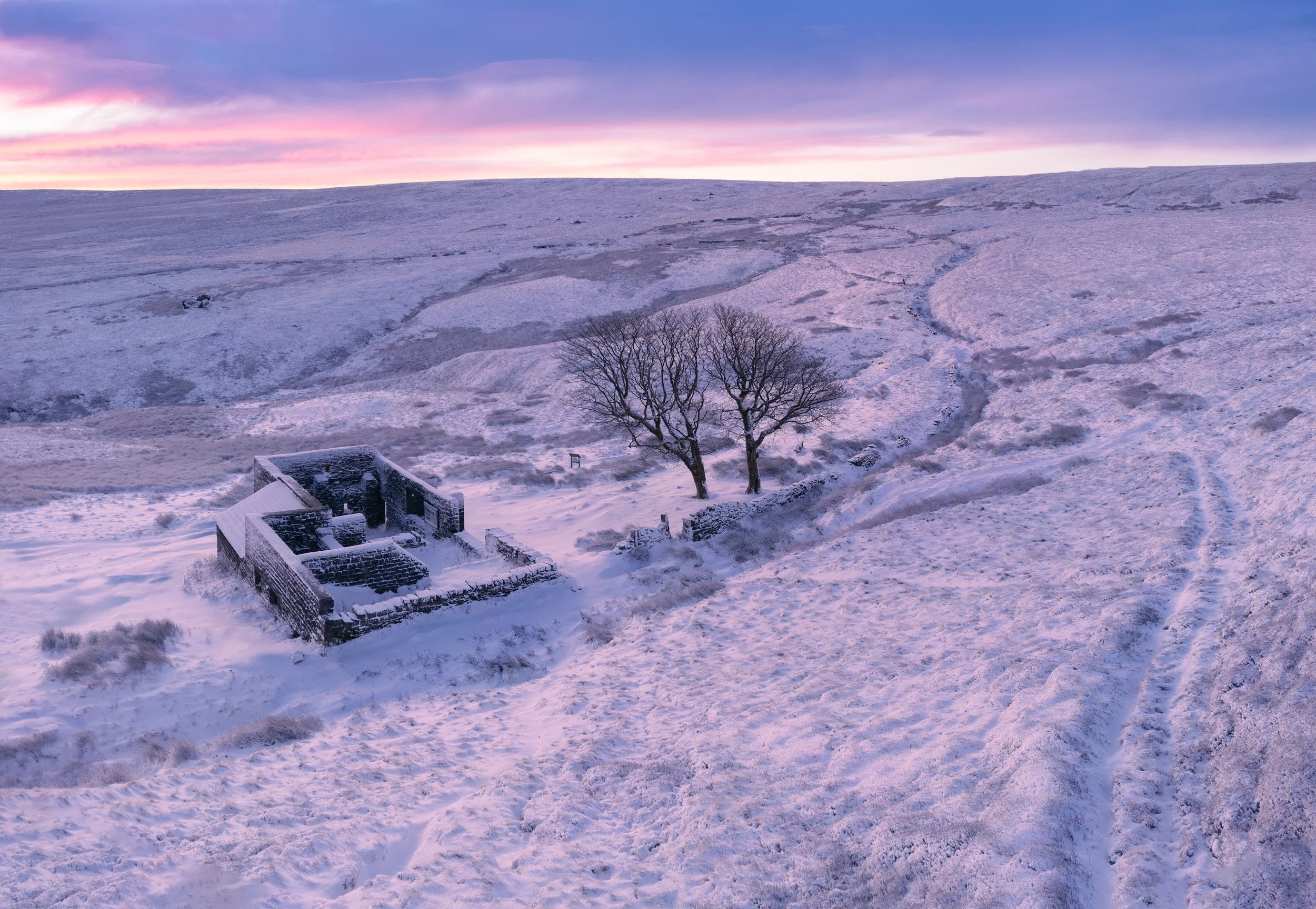 Snow-covered landscape during dusk with a pink and purple sky, a few leafless trees, and a small stone structure.