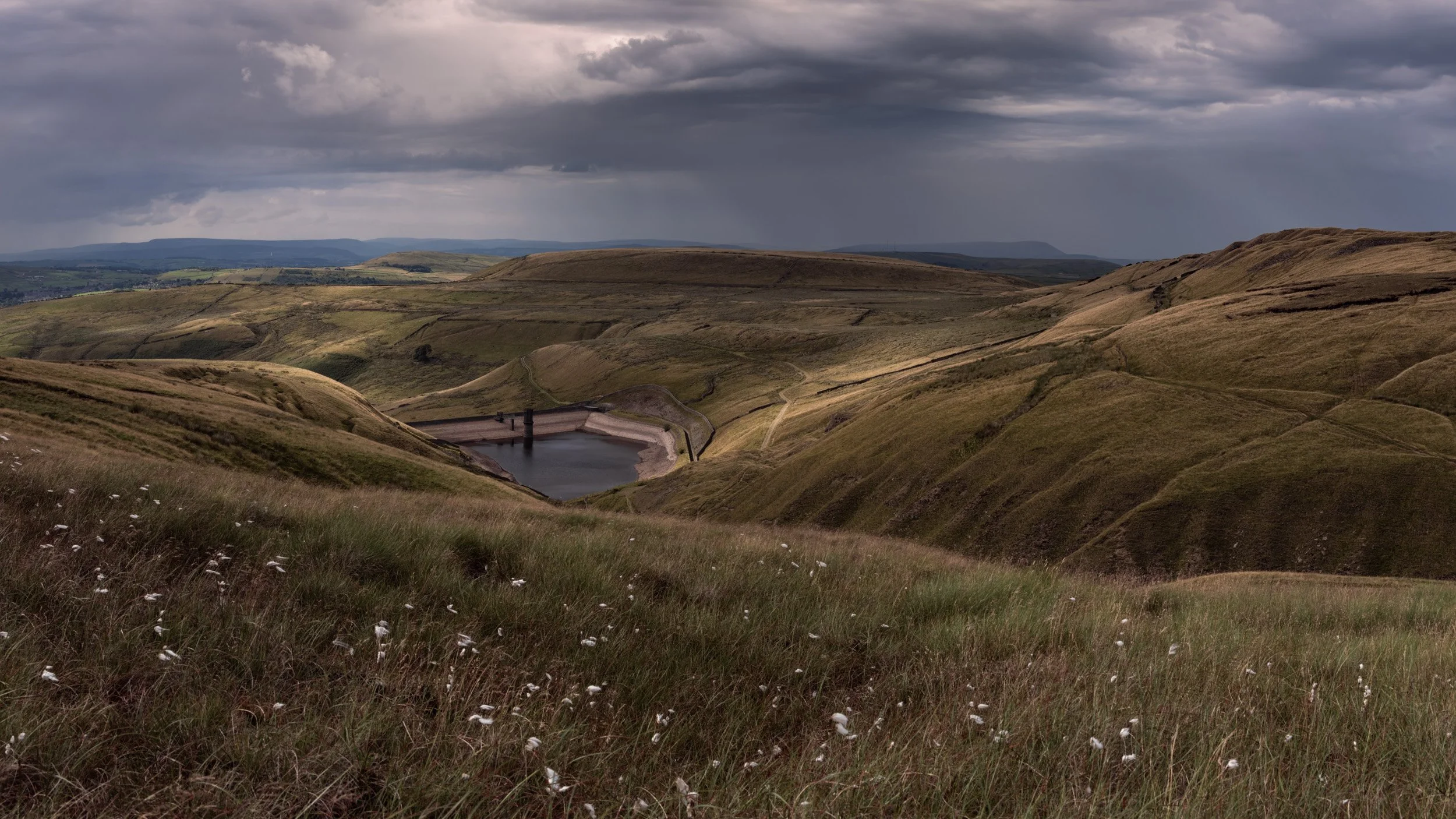 White bog cotton grass in the foreground looking down into the valley toward Scout Moor High Level Reservoir under dark storm clouds.