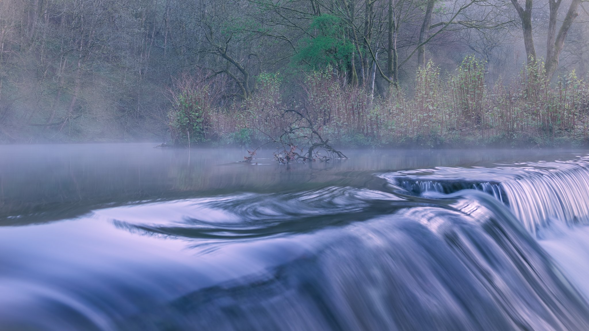 Weir on the River Roch in full flow with morning mist drifting over water in Rochdale