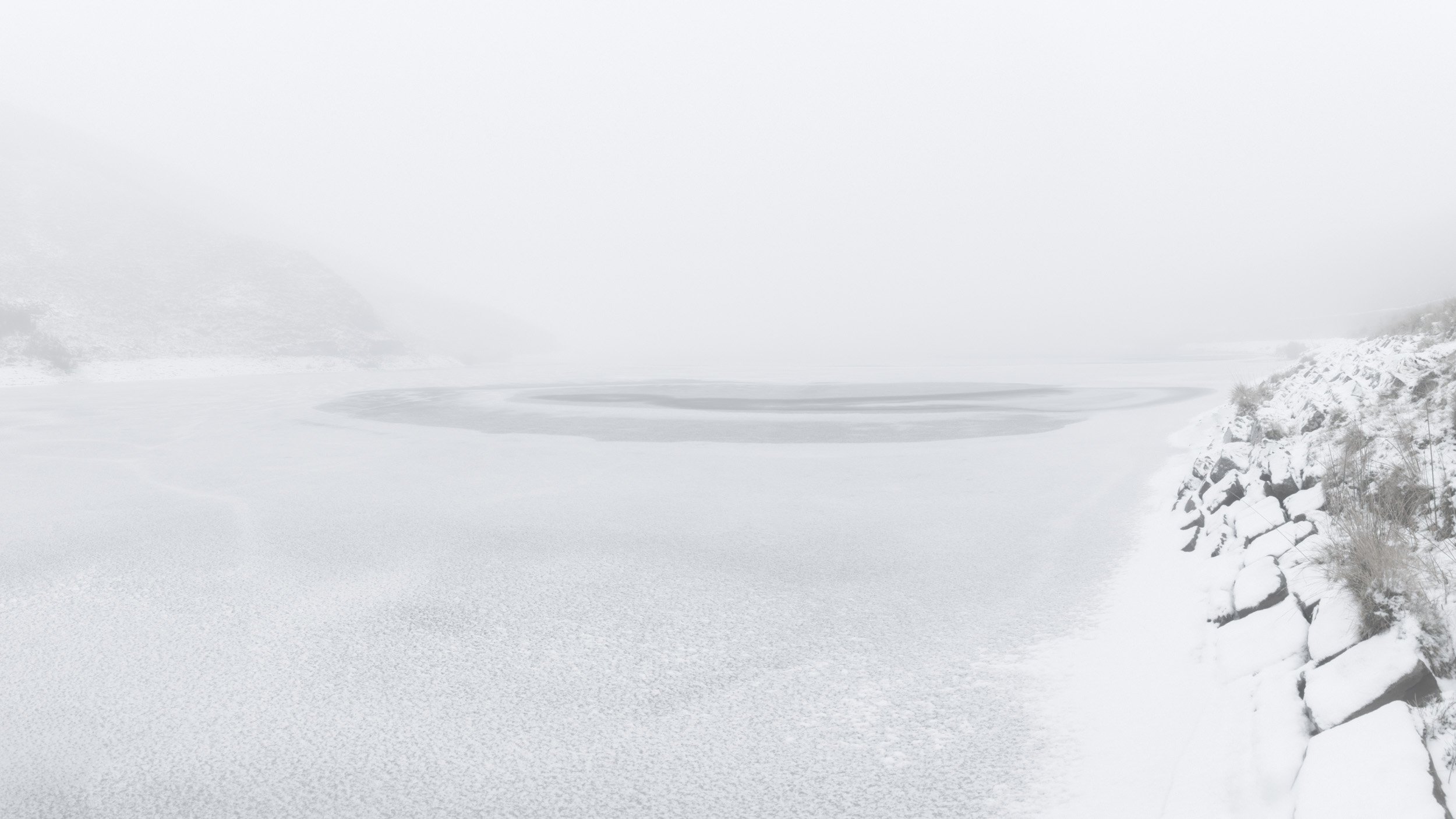 Minimalist high-key snow view of the frozen Scout Moor High Level Reservoir surrounded by dense fog and snow-draped rocks.