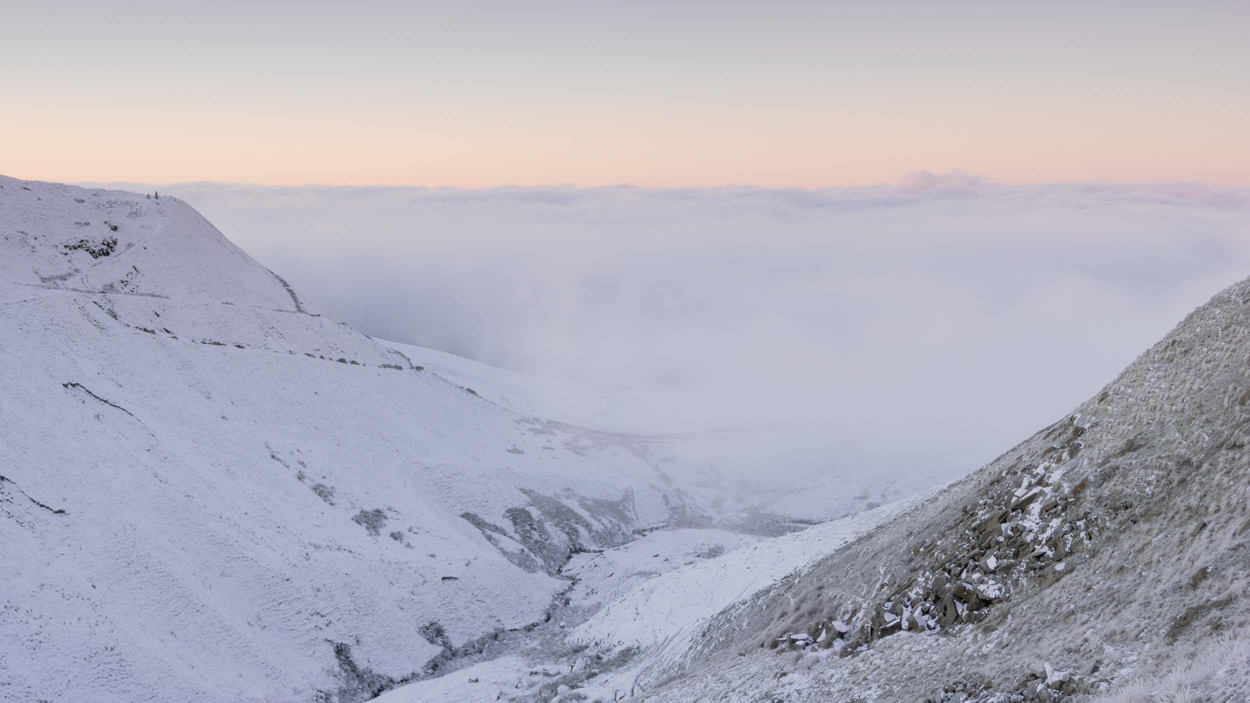Snow-covered steep slopes of Newgate Valley in Rossendale leading down to a valley filled with a dense cloud inversion under a soft pink sunset sky.