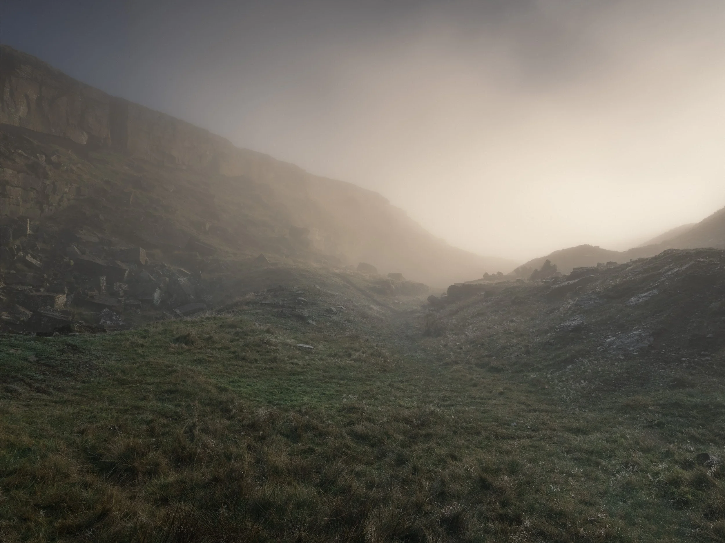 Grassy floor and towering stone quarry face of Fecit End Delf near Fecit Hill on Scout Moor, shrouded in soft, glowing fog.