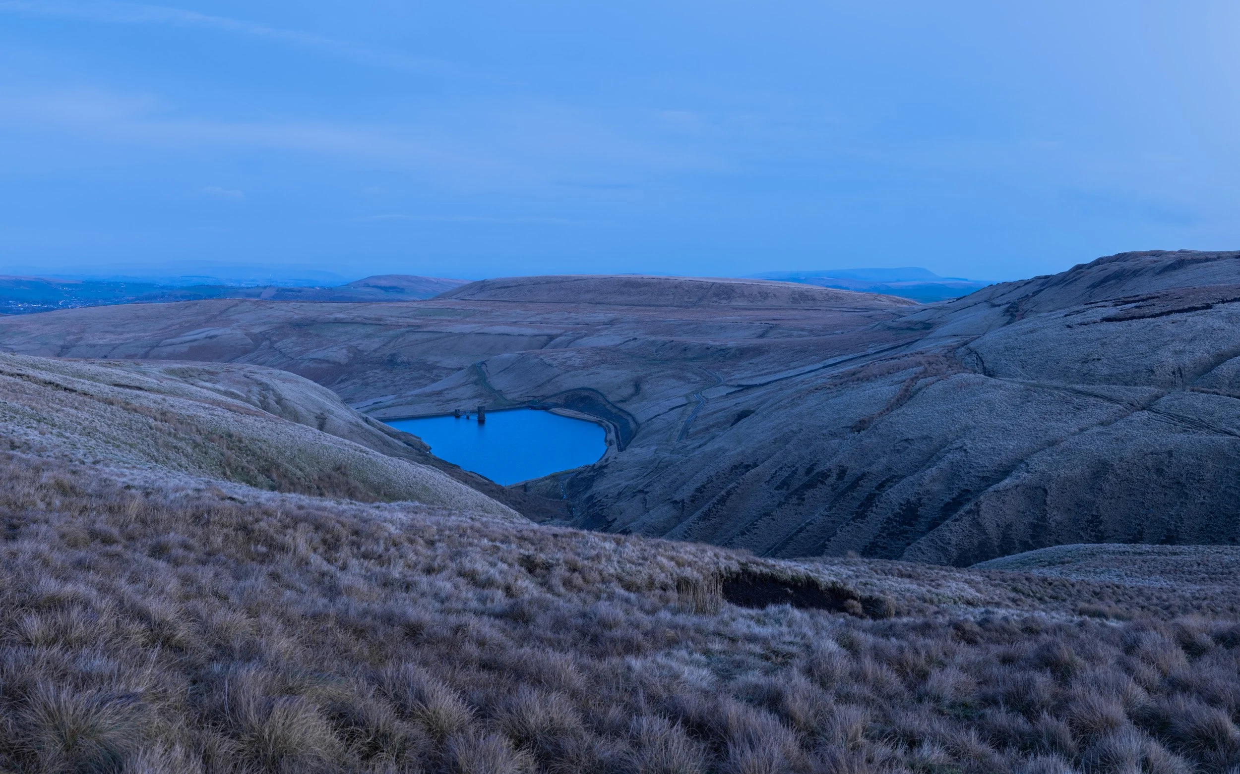 Deep blue hour landscape looking down onto Scout Moor High Level Reservoir nestled between frosted, rolling hills.