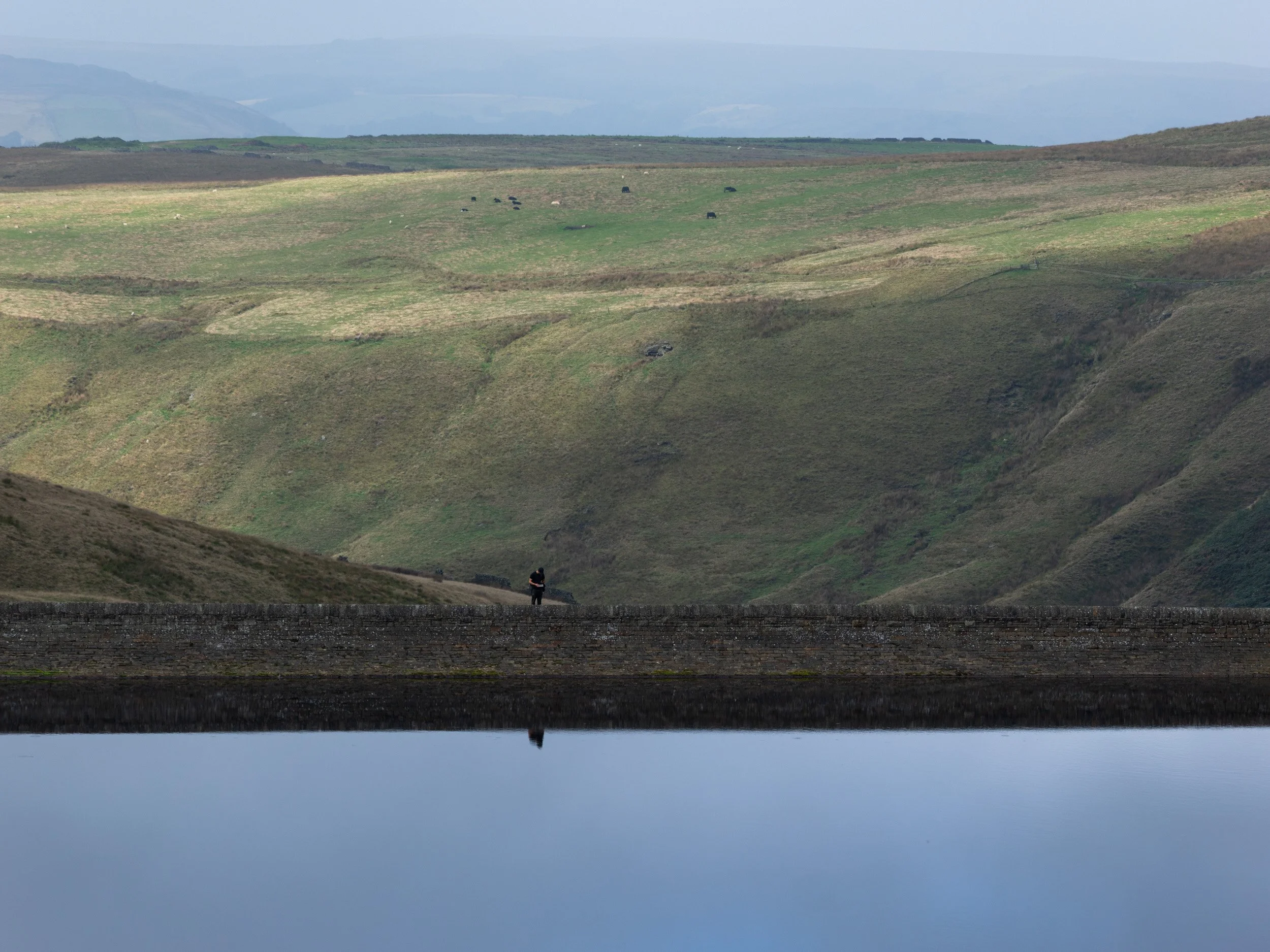 Silhouette of a lone walker standing on the stone embankment of Scout Moor High Level Reservoir reflected in still water.