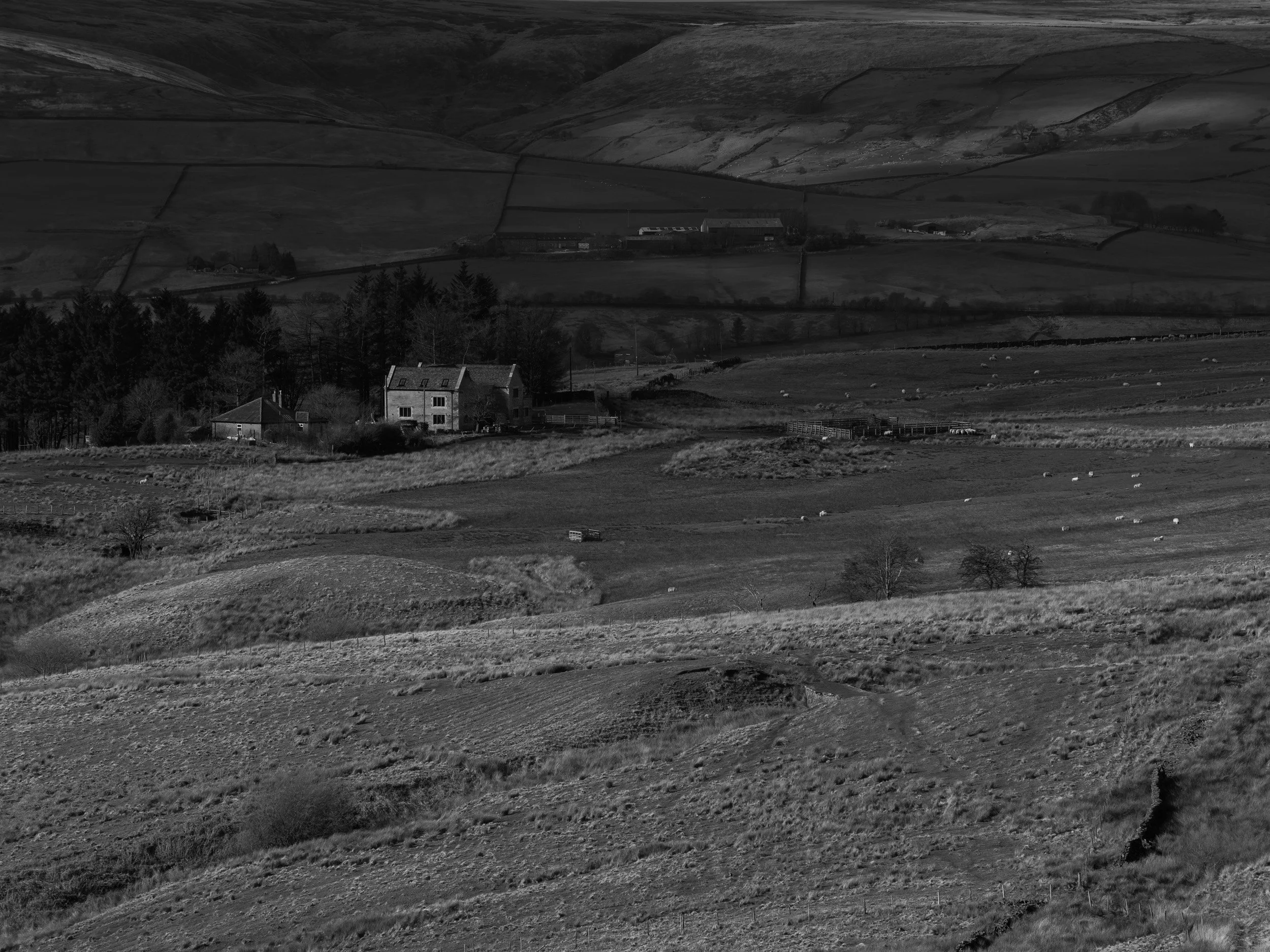 Black and white telephoto photograph looking down the slopes of Dearden Clough toward isolated stone farmhouses and drystone walled fields in Rossendale.